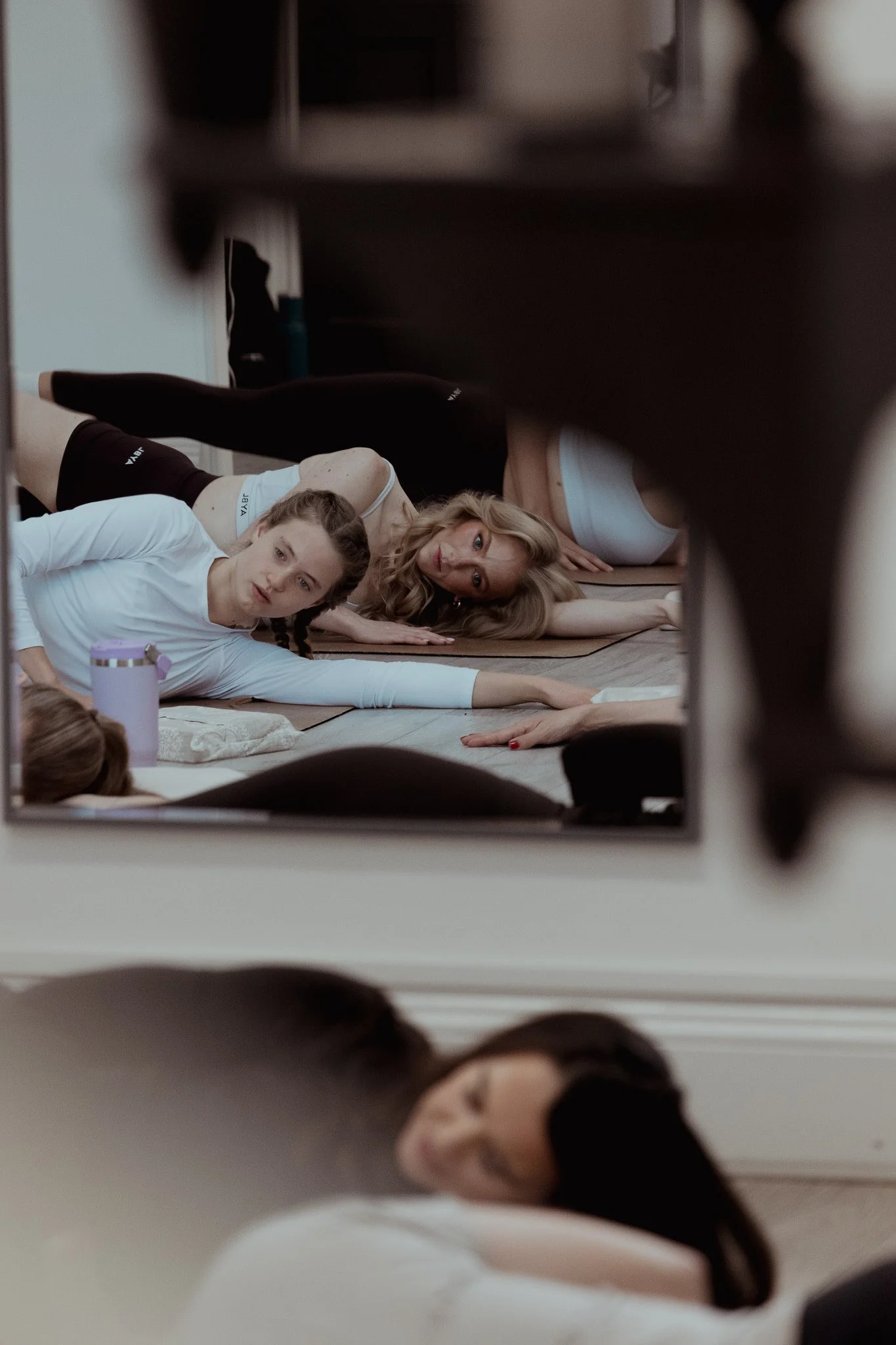 Two women practicing yoga on mats in a studio, viewed through a mirror, with a focus on their relaxed poses and focused expressions.