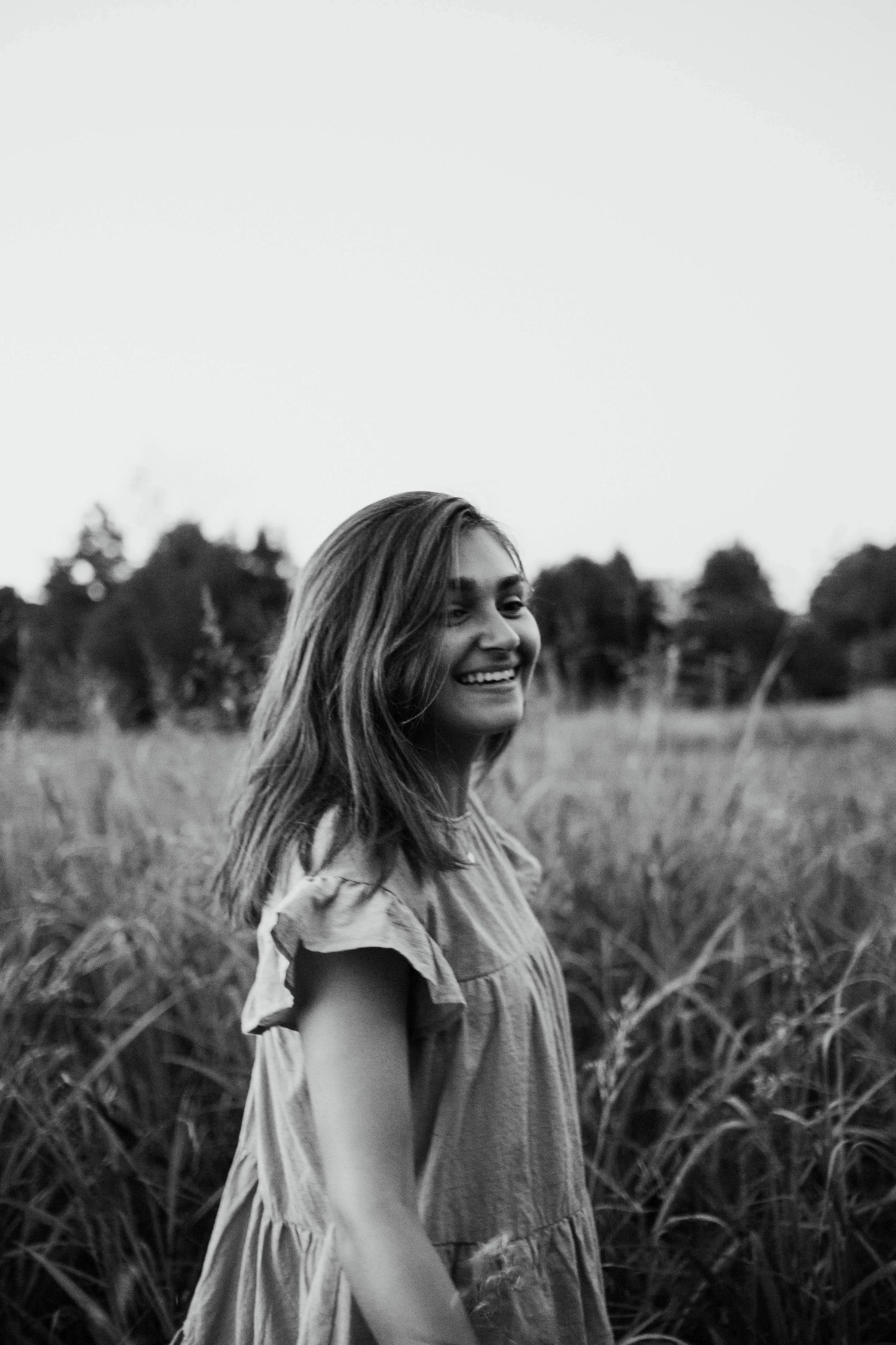 A young woman with shoulder-length hair smiling while standing in a field of tall grass, black and white photograph.