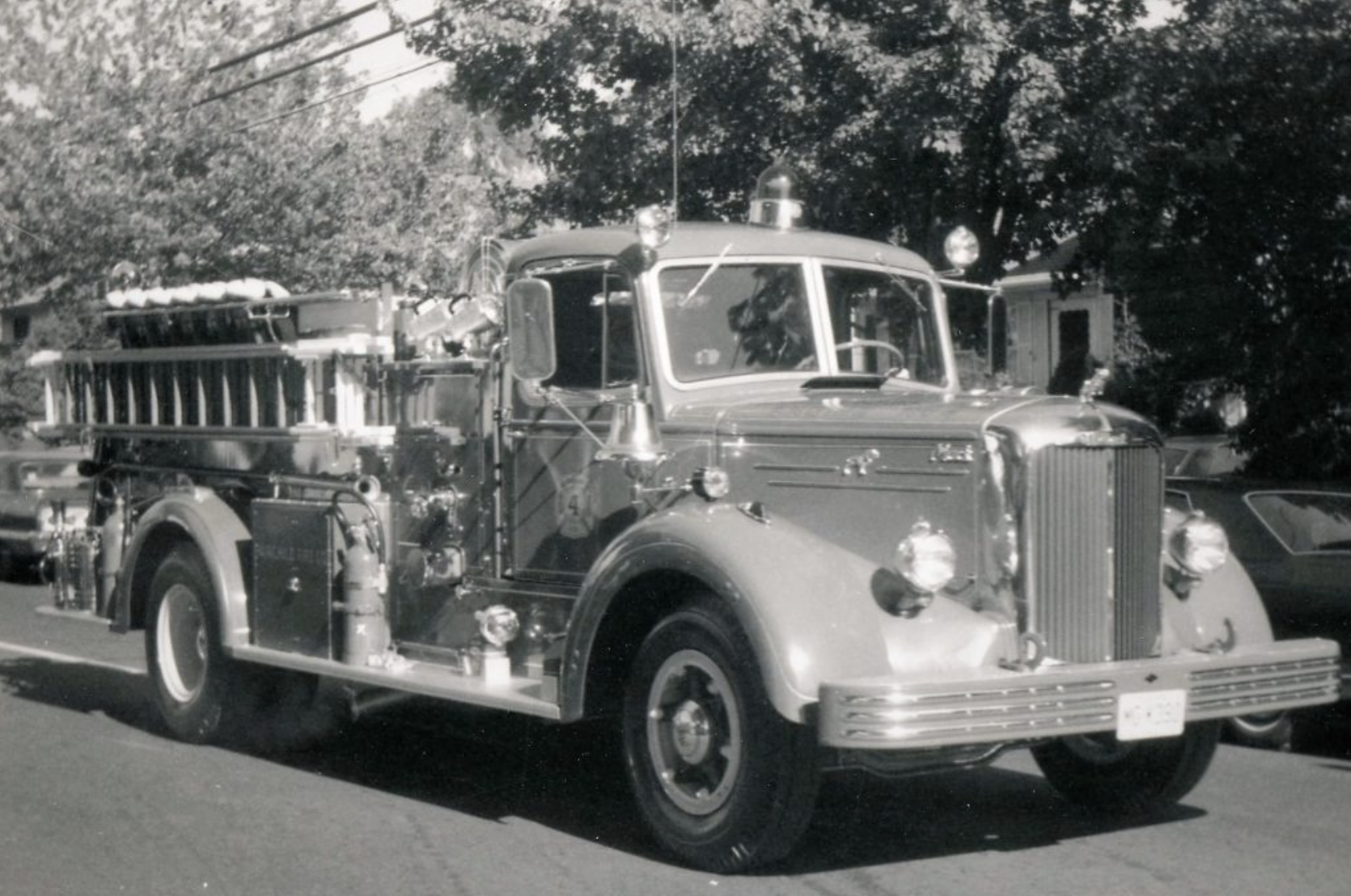 Black and white photo of a vintage fire truck parked on a street, with trees and other vehicles in the background.
