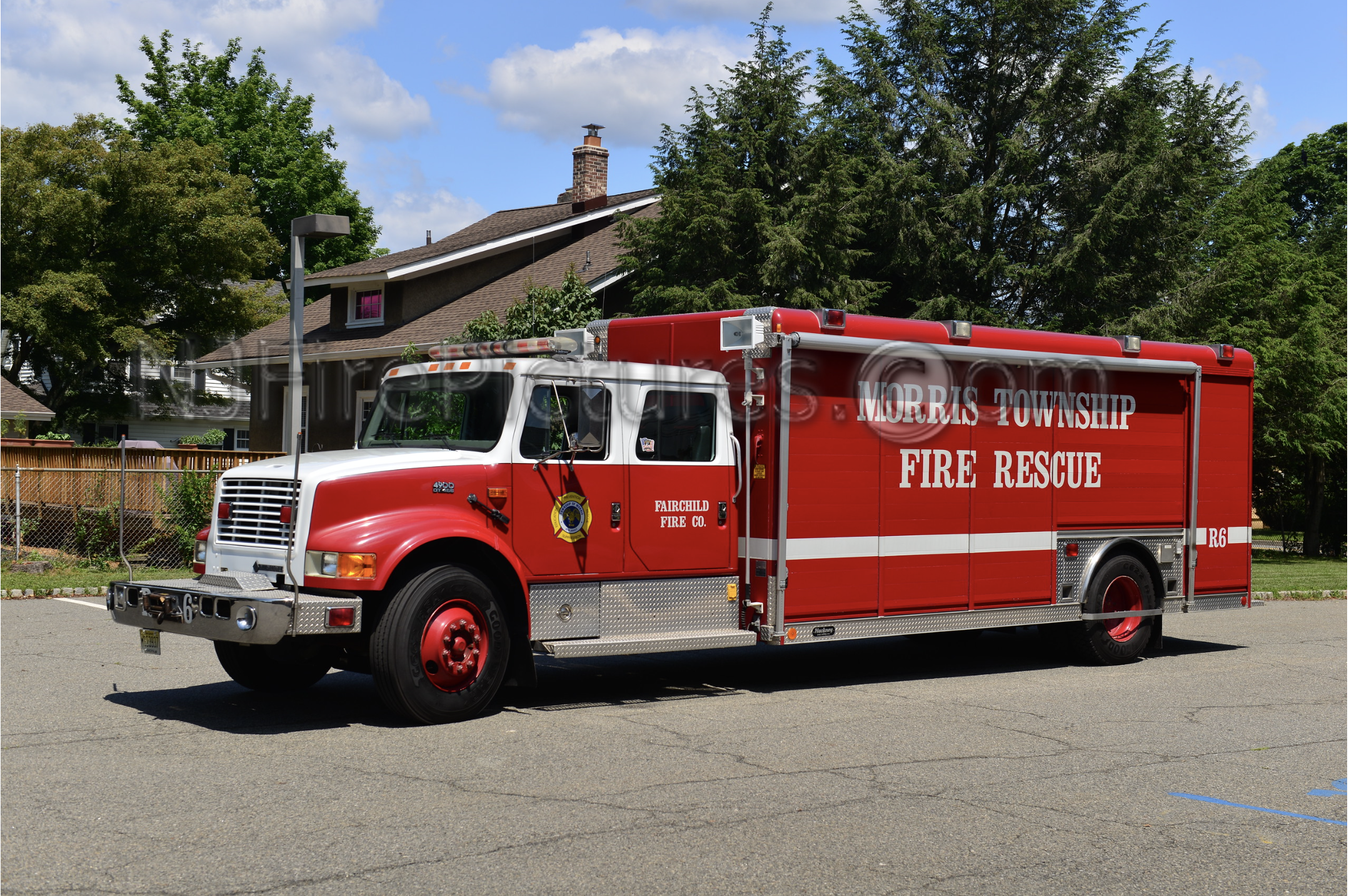 Red fire rescue truck with white lettering, parked on a paved area with trees and houses in the background.