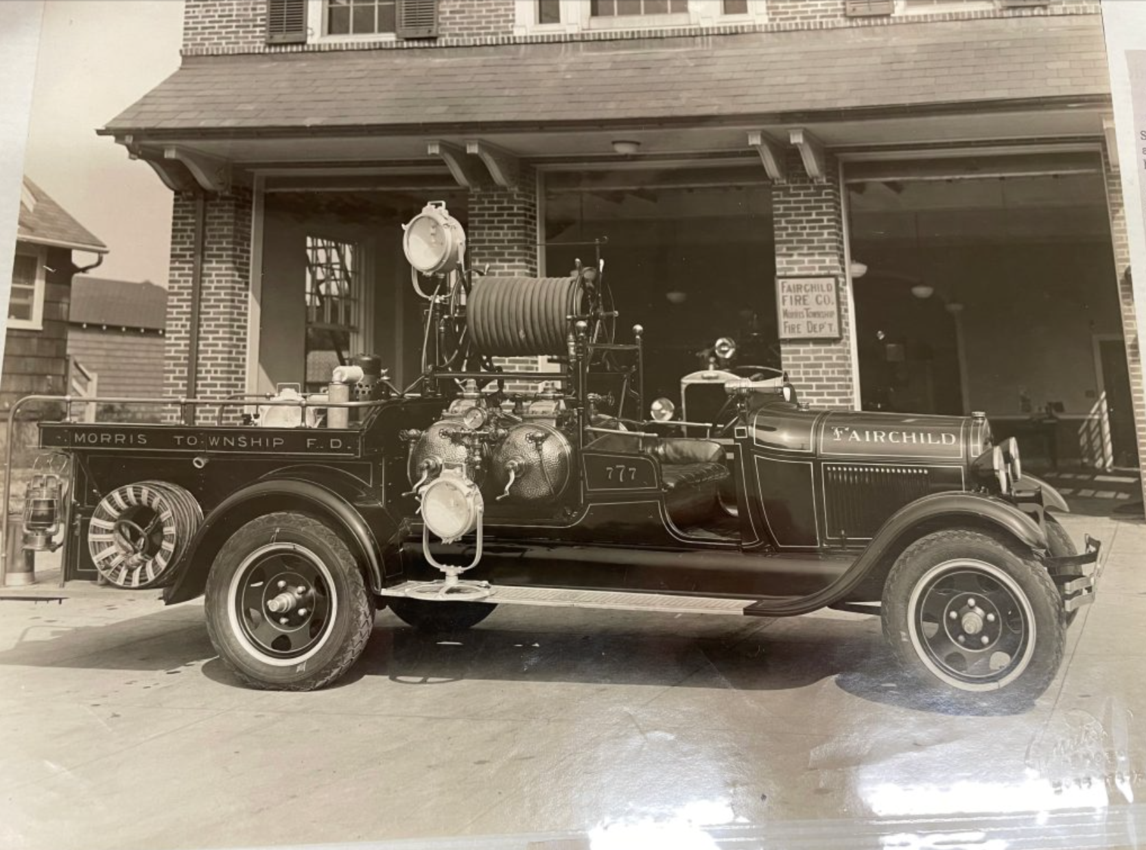 Historical fire truck parked outside a brick fire station, labeled 'Fairchild' and 'Morris Township Fire Department.'
