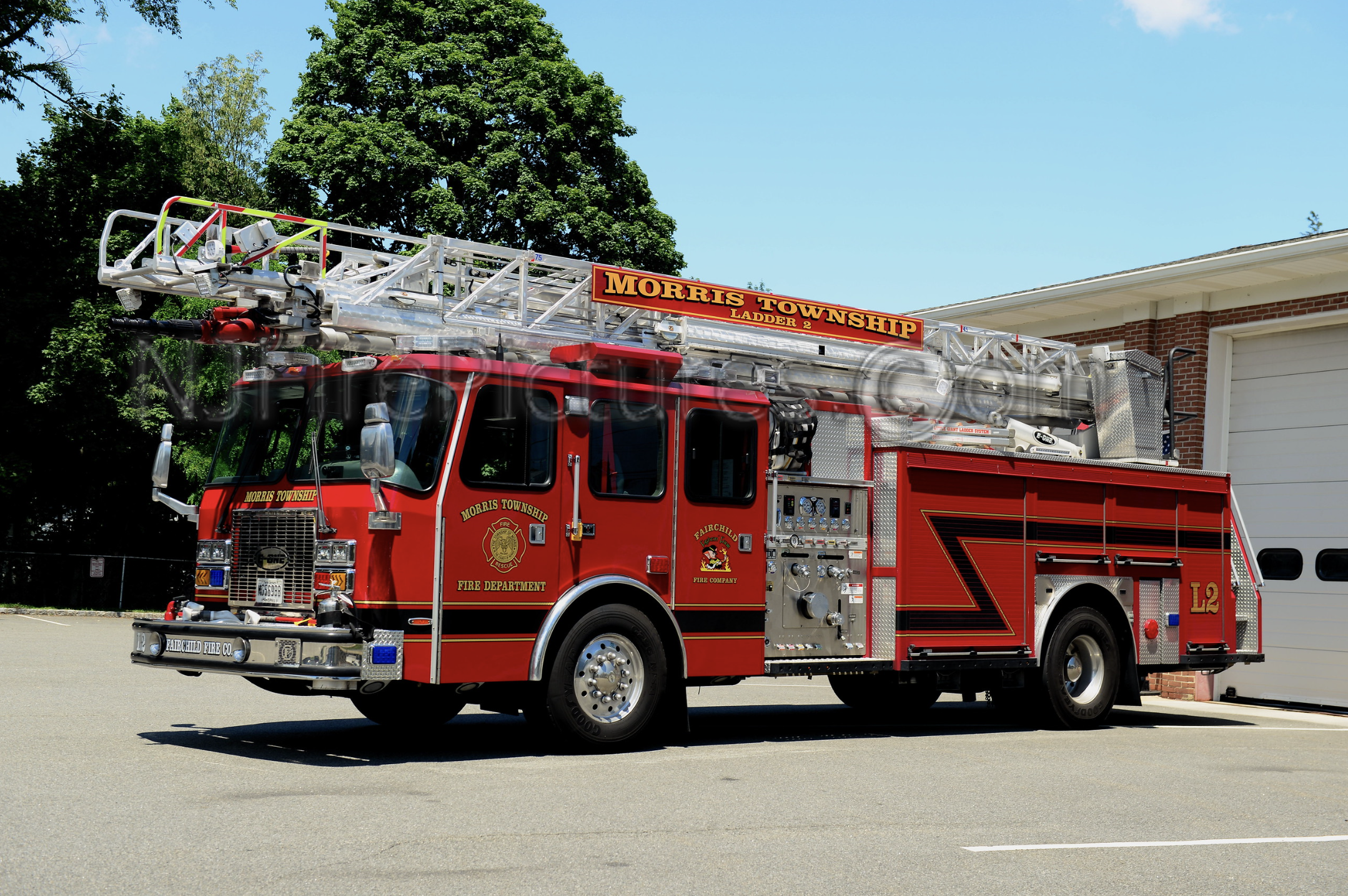 Red fire truck labeled 'Morris Township' with a ladder on top, parked on a street near a brick building and trees.