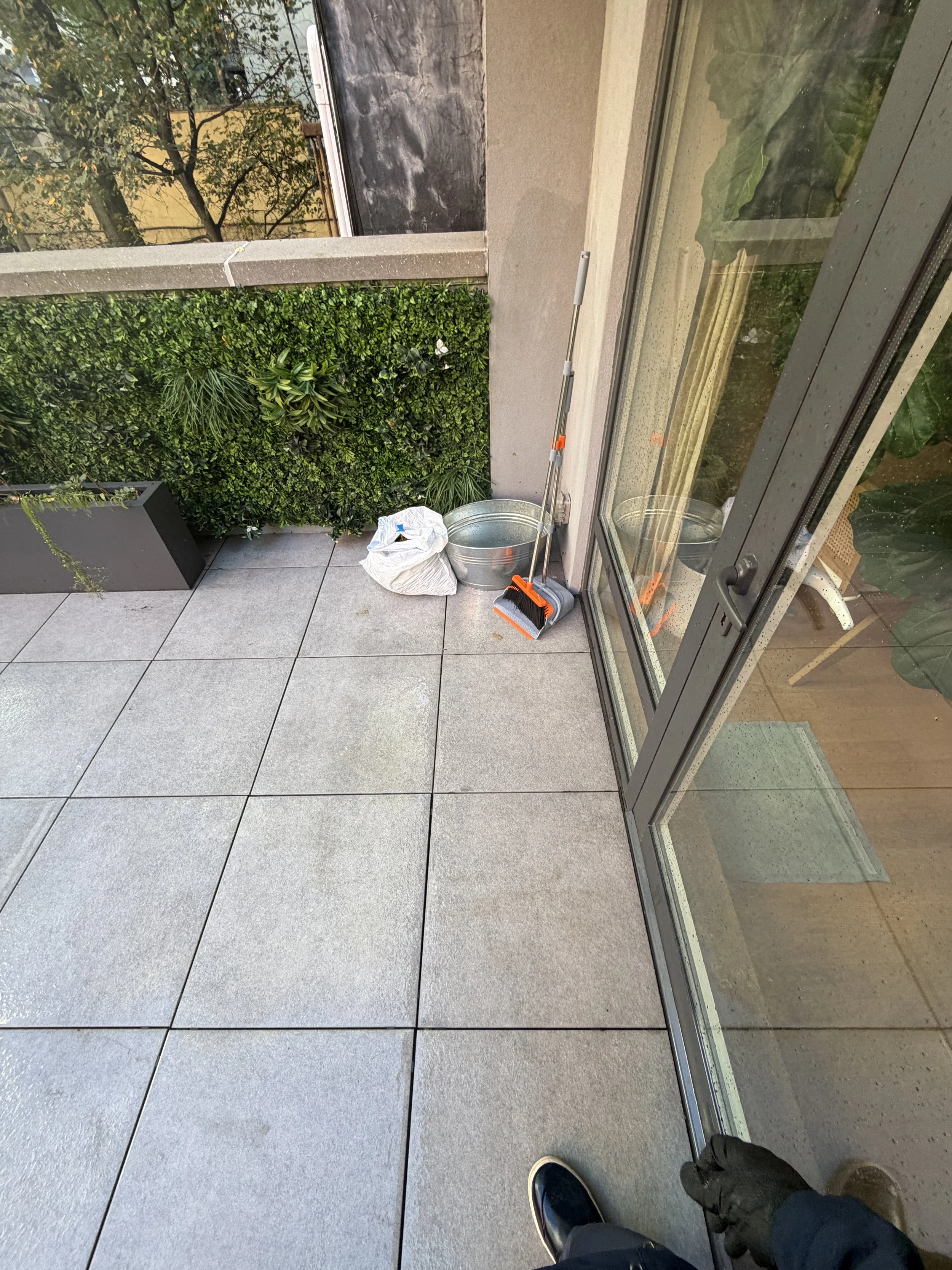 Terrace paving beside a glass door, with a broom, bucket, and bag gathered near the wall.