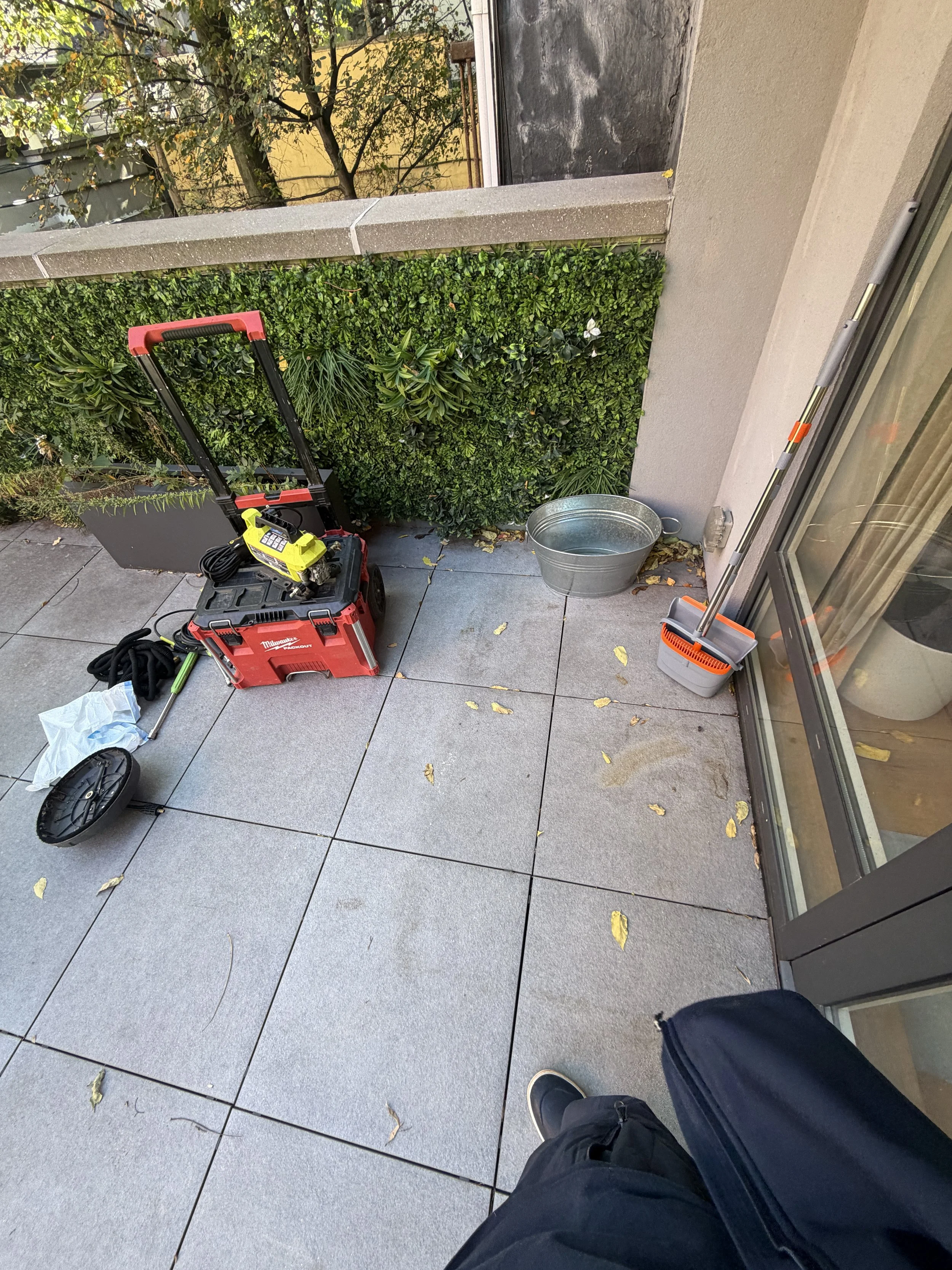 Leaves and surface marks on terrace paving near a glass door, with cleaning equipment nearby.
