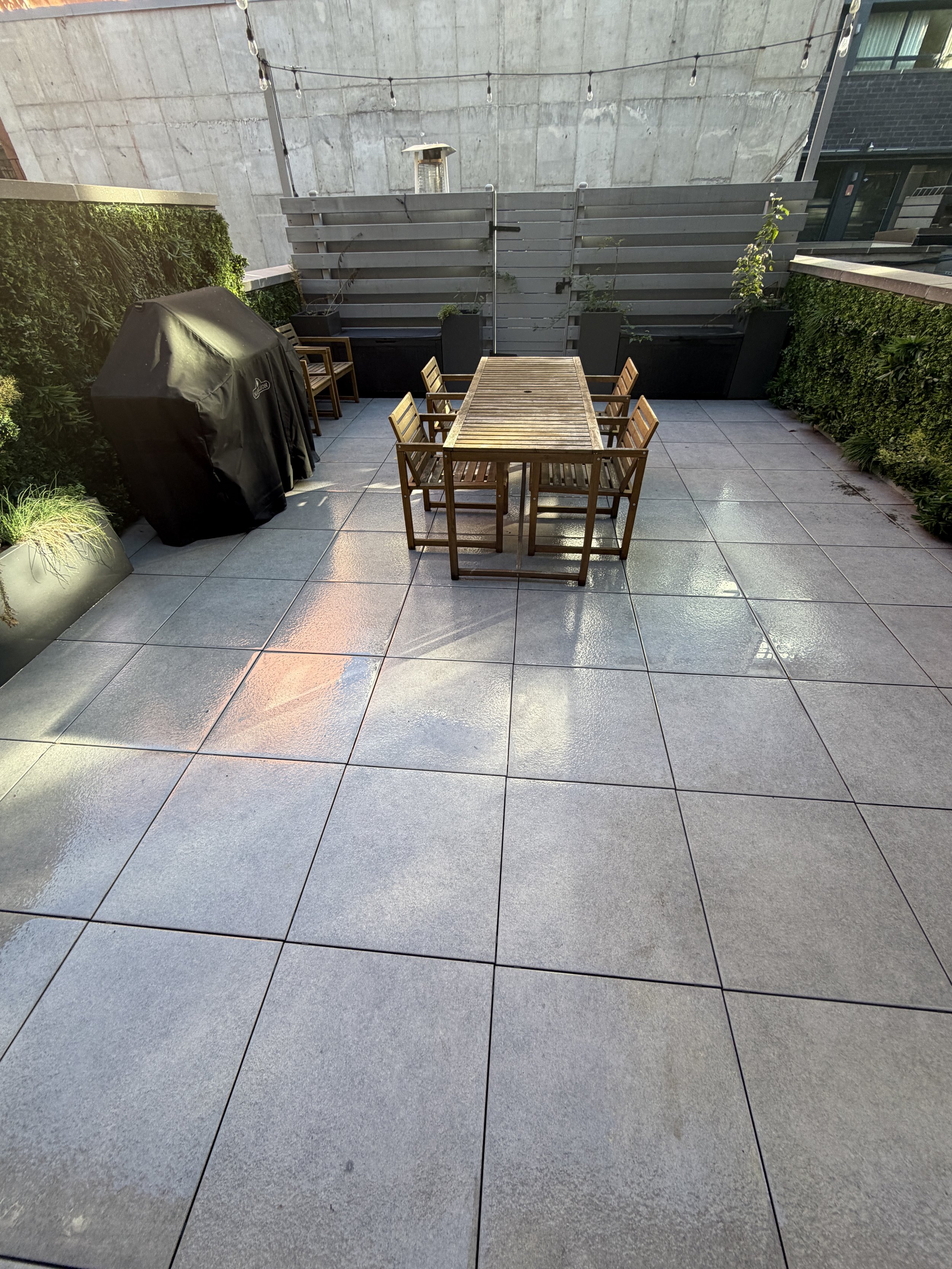 Wet gray terrace paving surrounding a wooden dining table and chairs, with a covered grill nearby.