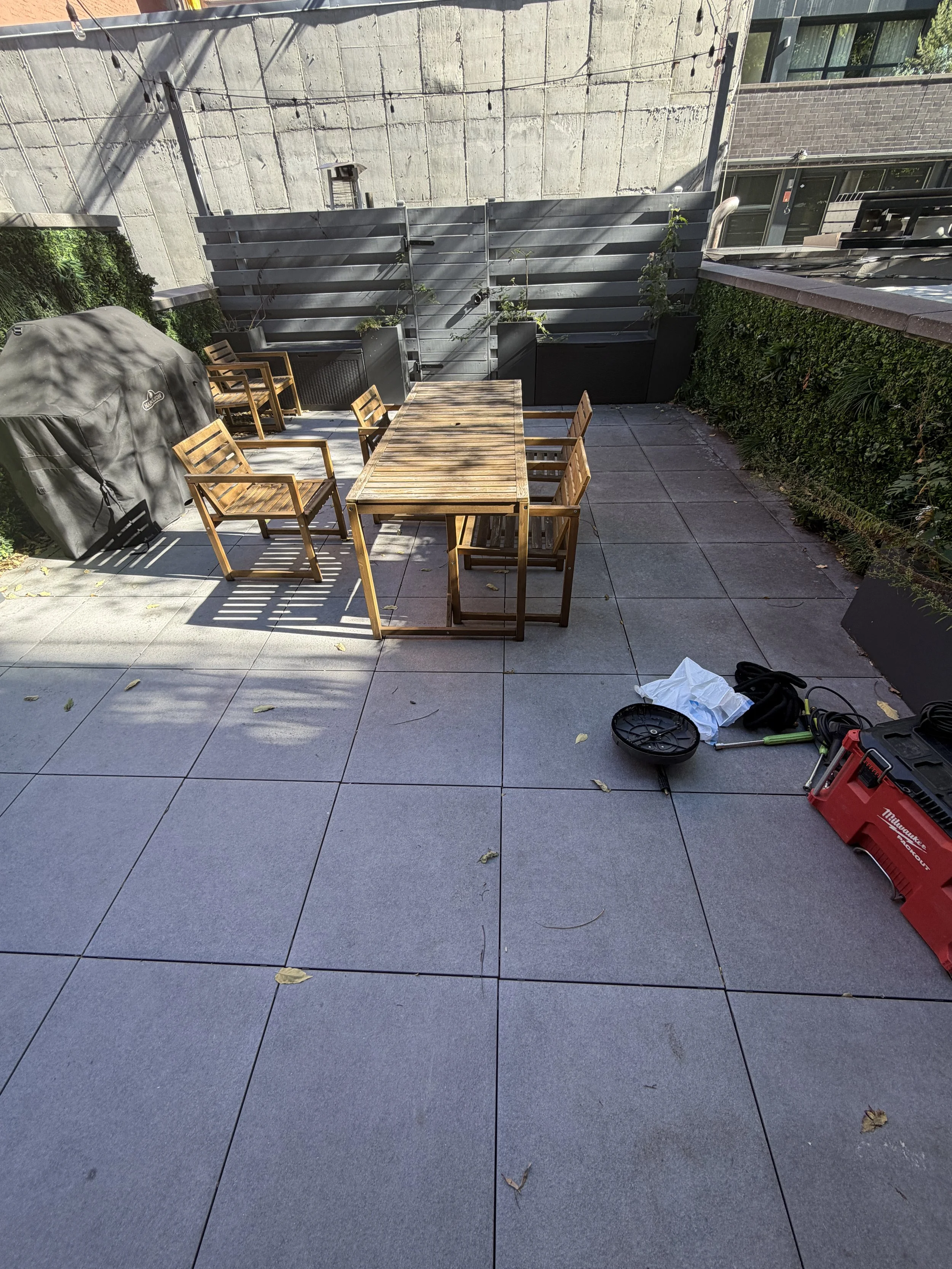 Gray terrace paving with scattered leaves around a wooden dining table and chairs, with a covered grill and cleaning equipment nearby.