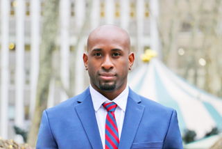 A man in a blue suit with a red and blue striped tie standing outdoors in front of a tent and trees.