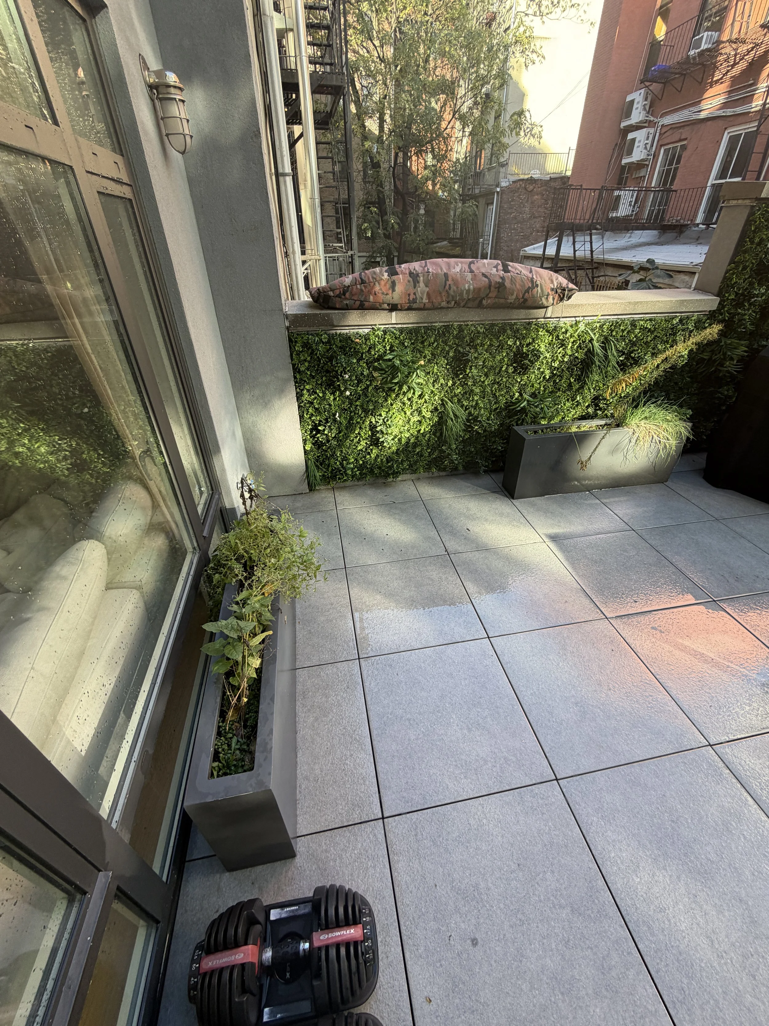 Wet terrace paving beside rectangular planters, large windows, and a green privacy screen.