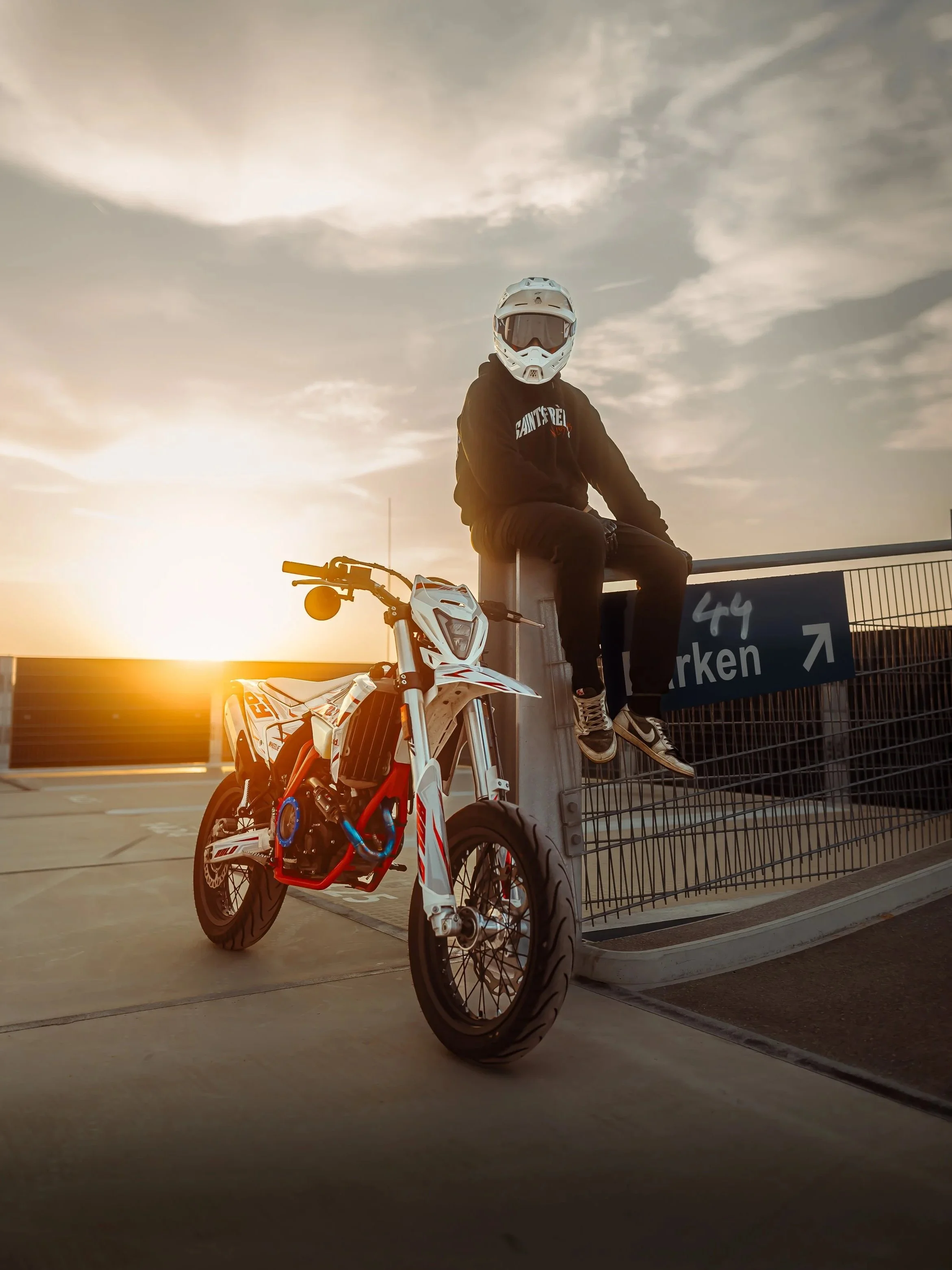 Person in Motorradhelm und dunkler Kleidung sitzt auf einer Mauer an einem Parkplatz mit einem weißen Motocross-Bike während des Sonnenuntergangs.