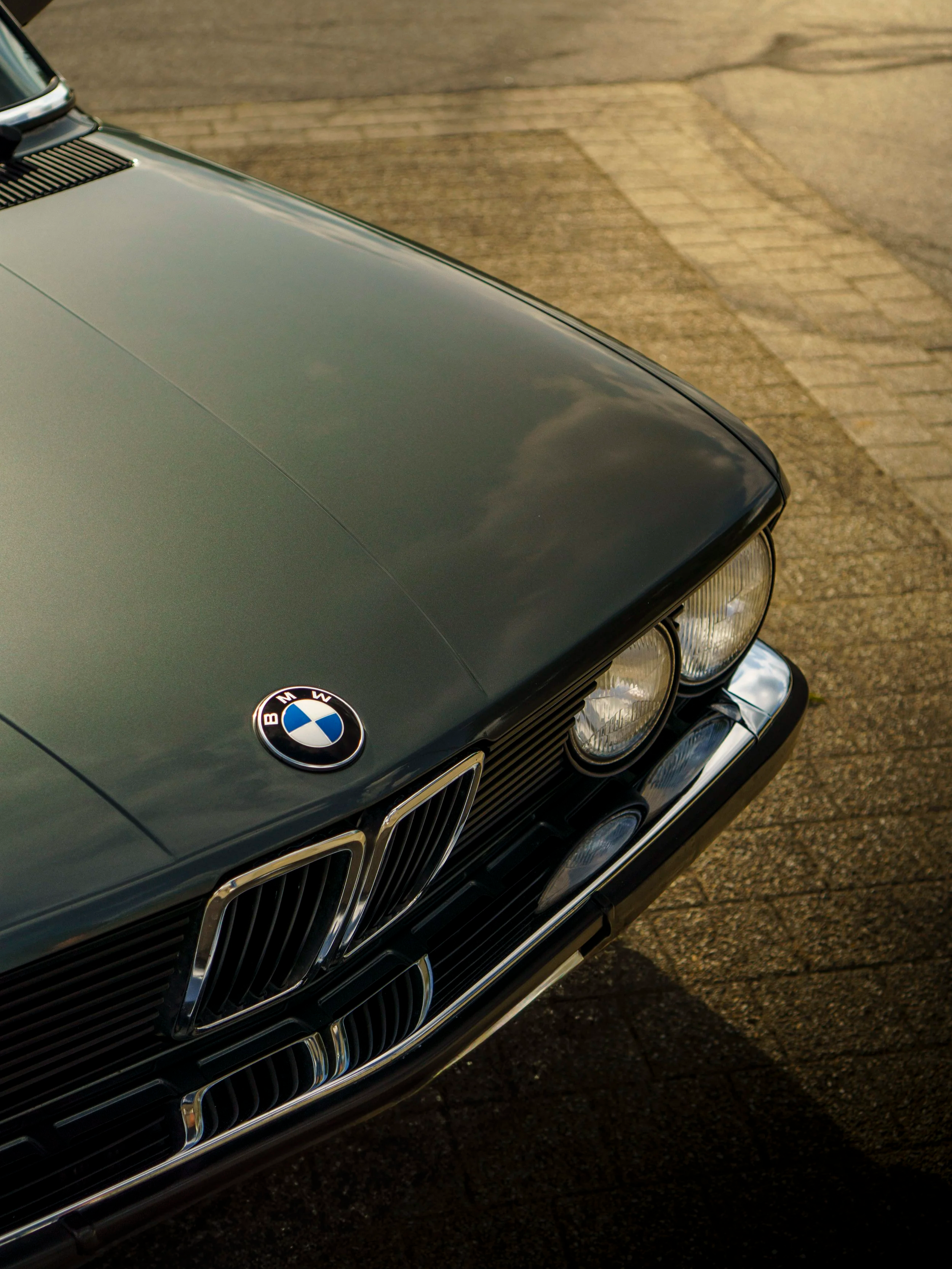 Nahaufnahme der vorderen Fahrzeugpartie eines BMW Autos, inklusive BMW-Logo, Scheinwerfer und Kühlergrill, auf einer asphaltierenden Straße bei Sonnenuntergang.