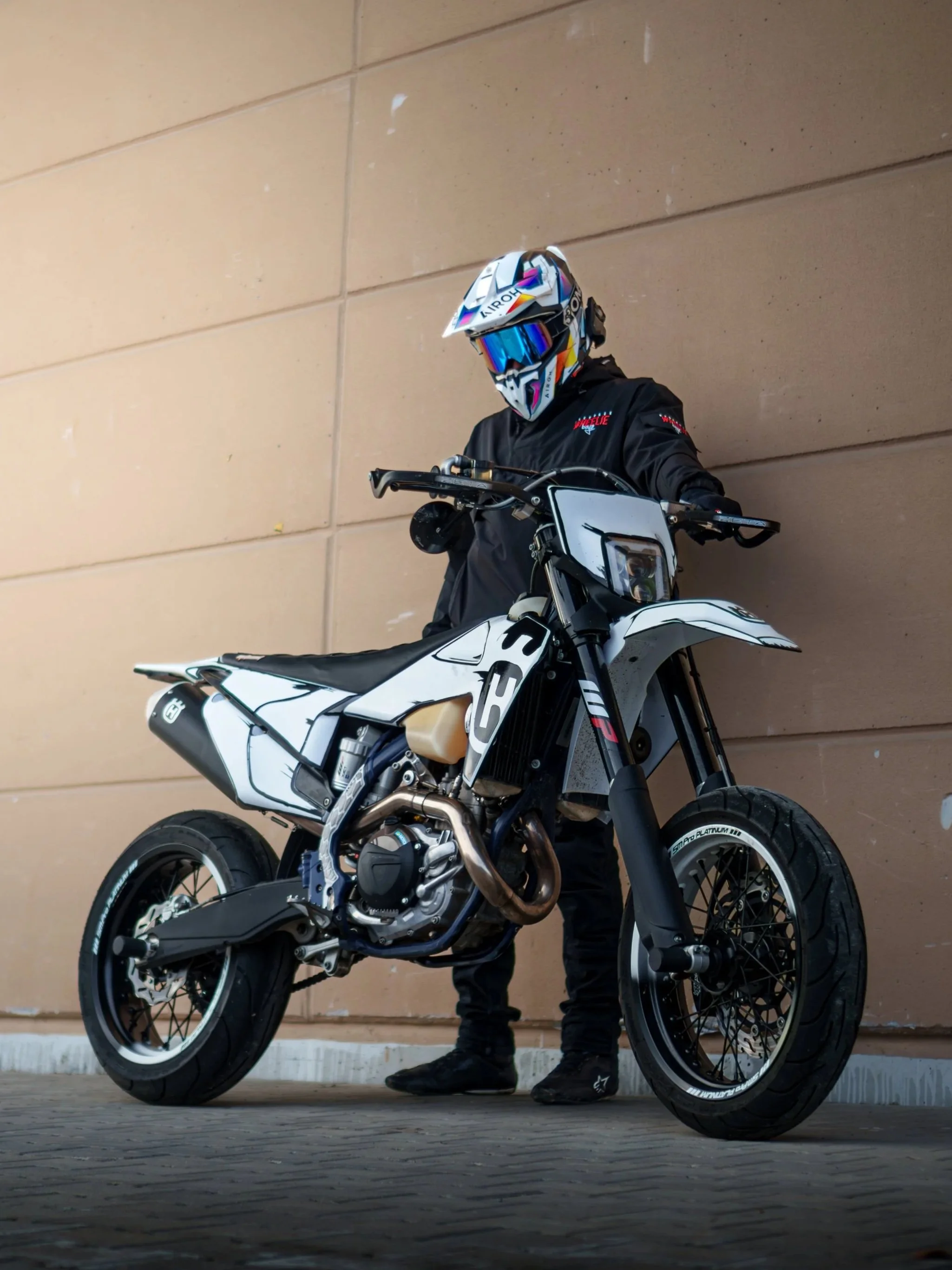 Person mit Motorrad, im schwarzen Motorradanzug und Helm, lehnt an eine braune Wand.