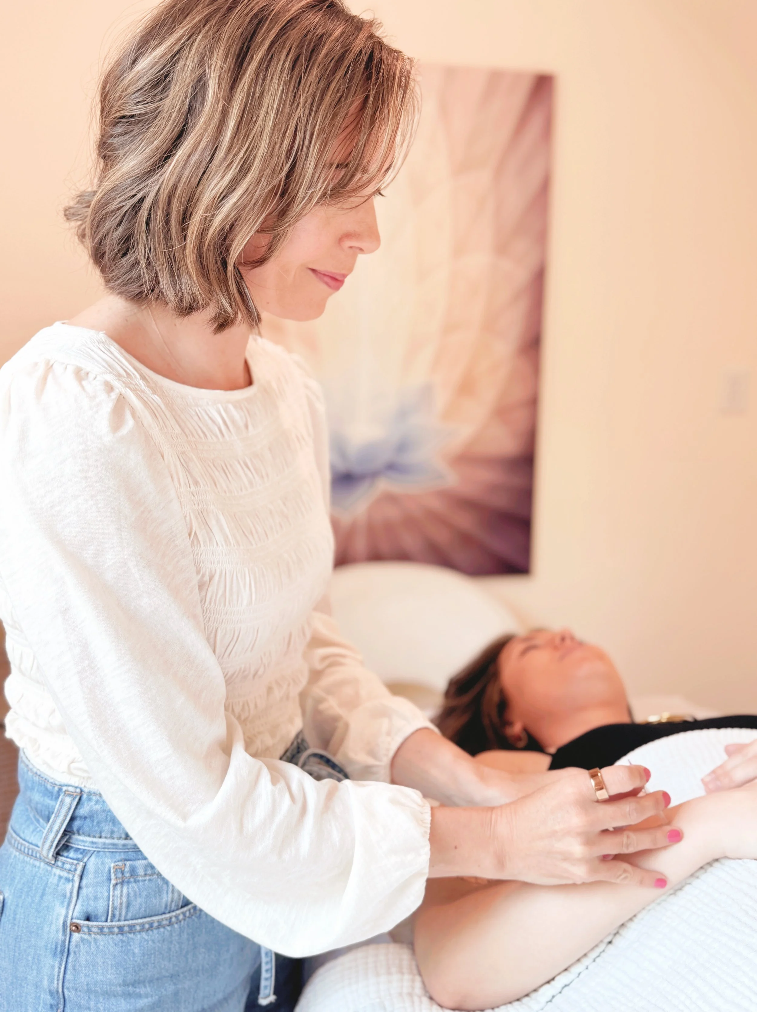 A woman with shoulder-length blonde hair in a white blouse is performing acupuncture on a woman lying down with her eyes closed, in a spa or beauty clinic setting.