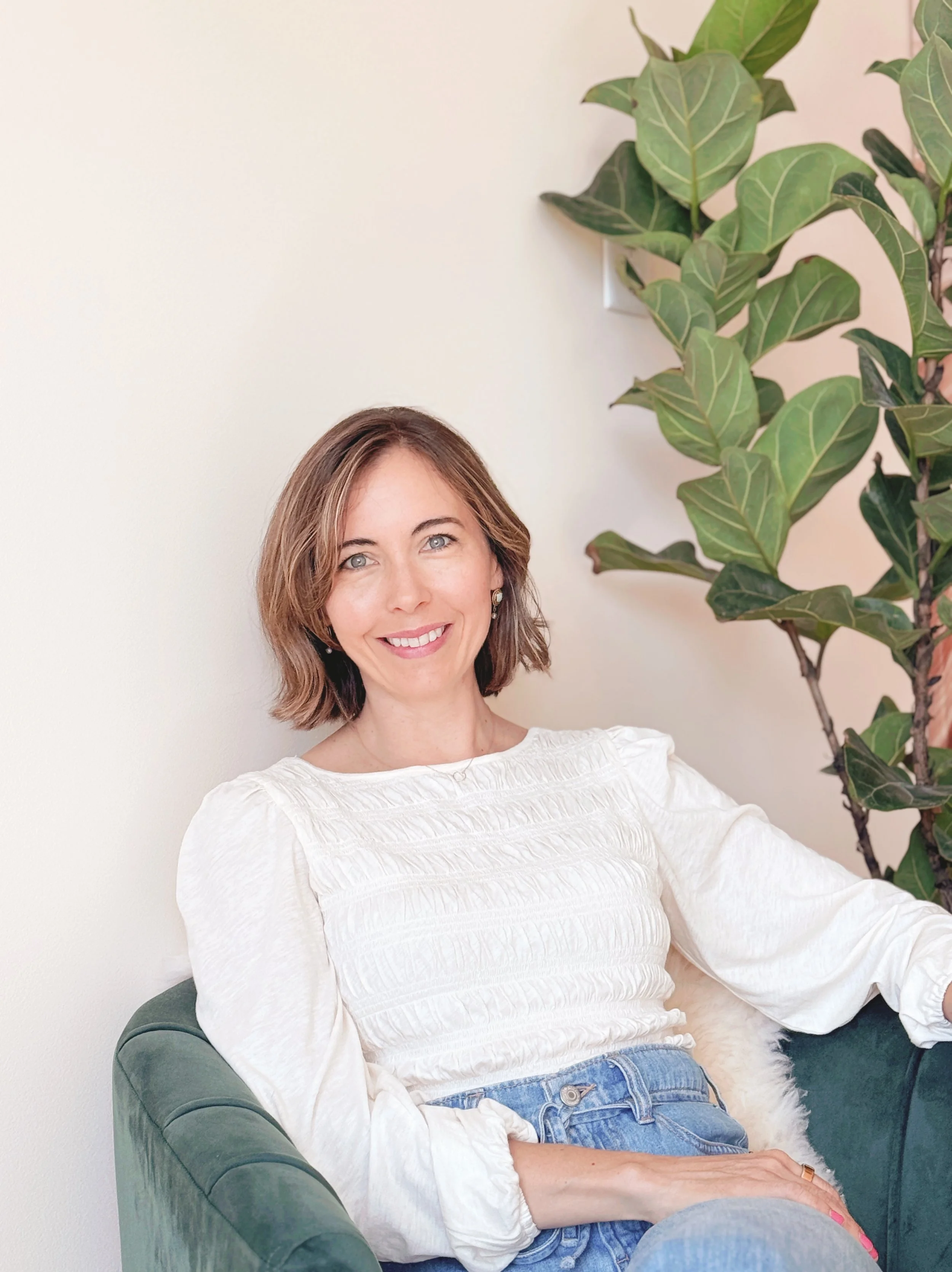 A woman with shoulder-length brown hair and blue eyes smiling, sitting on a green velvet armchair next to a large ficus plant with broad green leaves, wearing a white textured blouse and blue jeans.