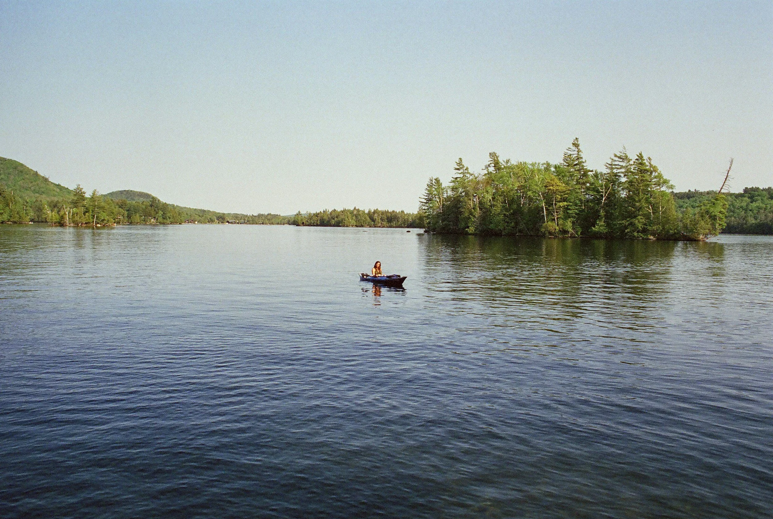 A person in a small boat on a calm lake with a small, tree-covered island nearby and forested hills in the background on a clear day.