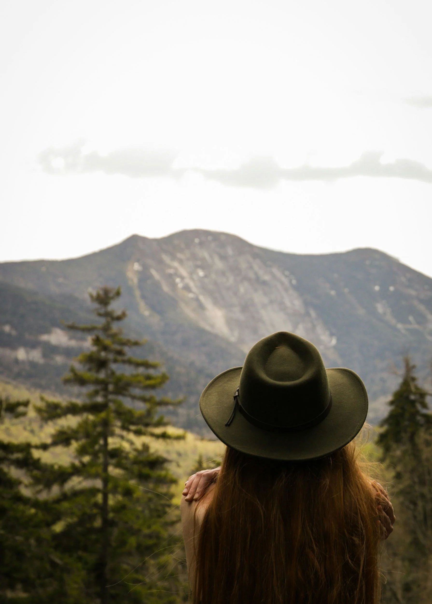 A woman with long hair wearing a wide-brimmed hat, sitting outdoors and looking at a mountain landscape with trees.