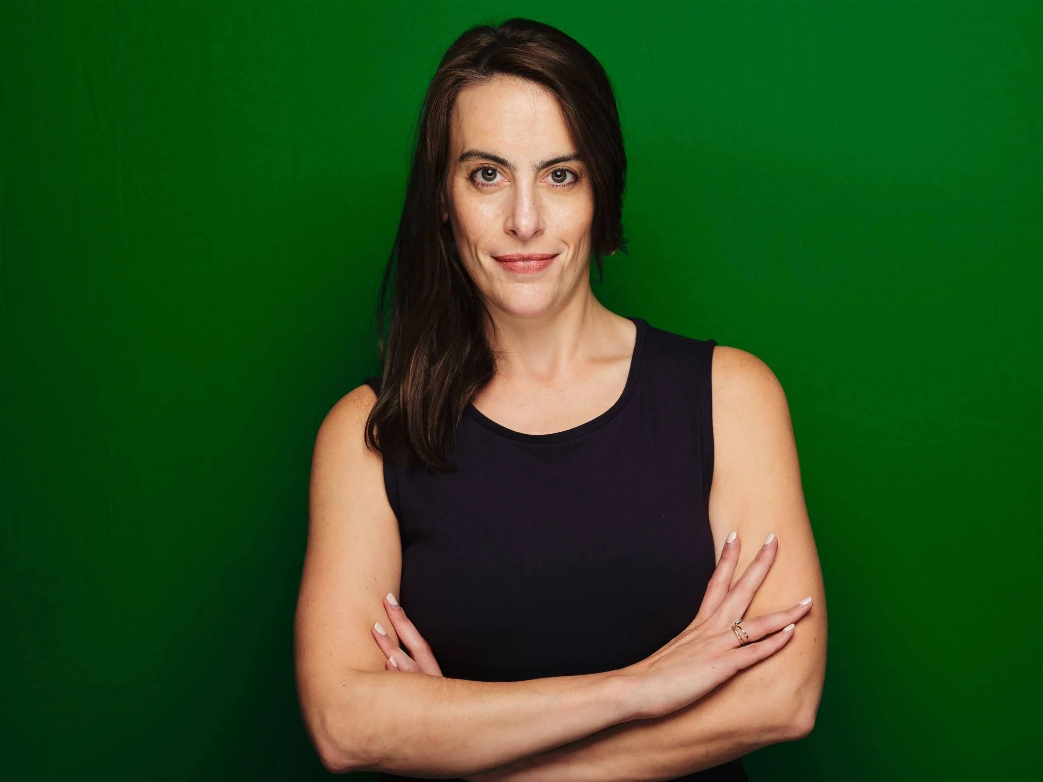 A woman with dark brown hair wearing a black sleeveless top standing with her arms crossed against a plain green background.