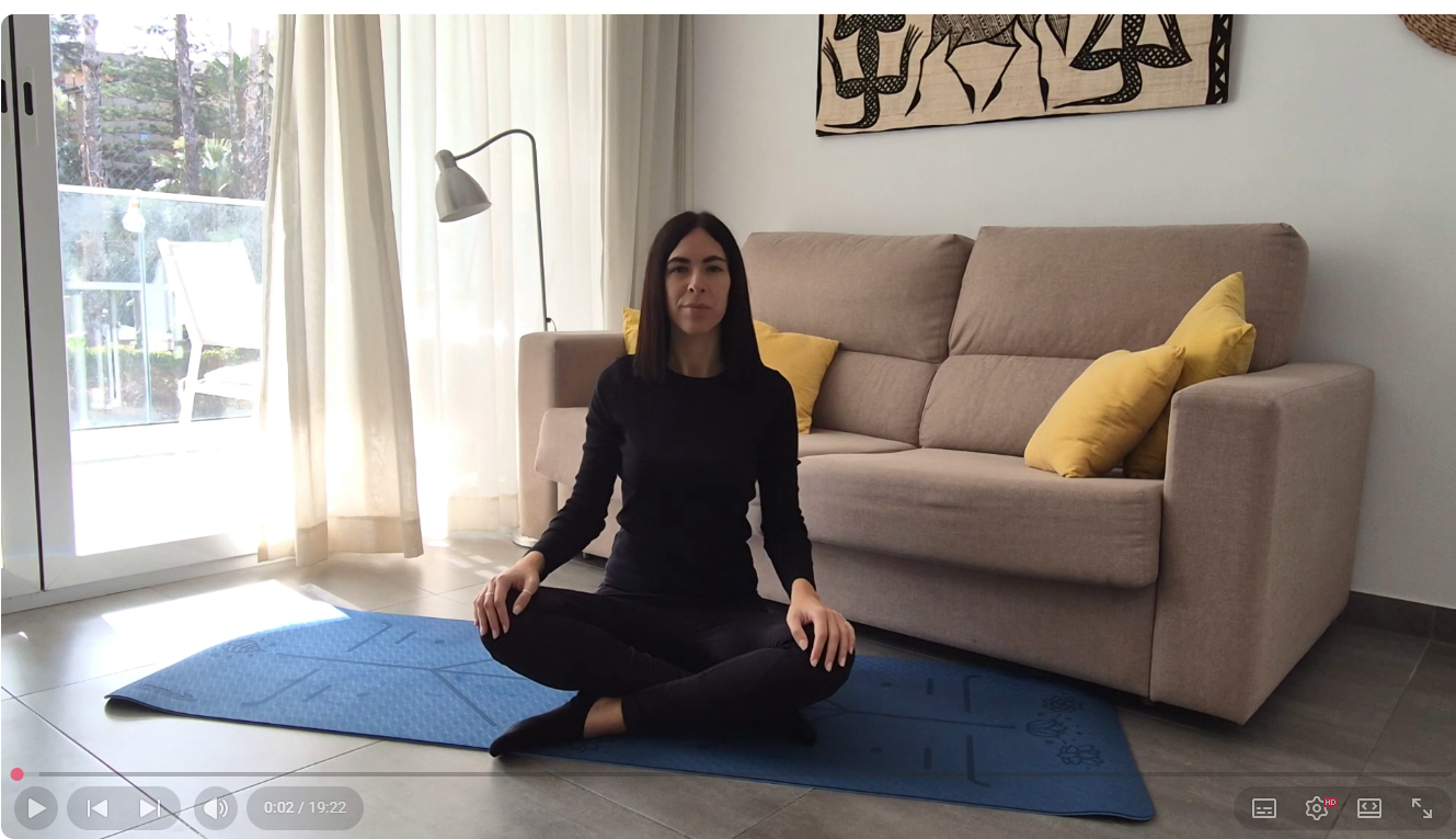 Una donna asiatica con i capelli neri, vestita di nero, seduta in posizione di meditazione sul pavimento con un tappetino da yoga blu, in soggiorno con divano beige e cuscini gialli, e molto naturale luce proveniente da una porta finestra.