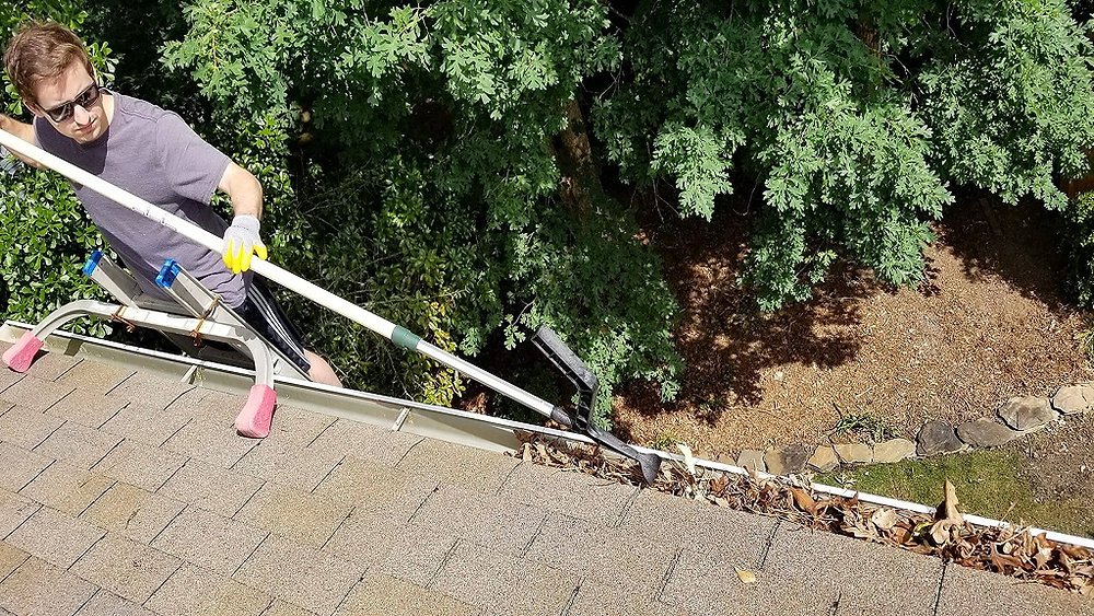 A person on a ladder is cleaning leaves and debris from a gutter on a residential roof using a long-handled tool.