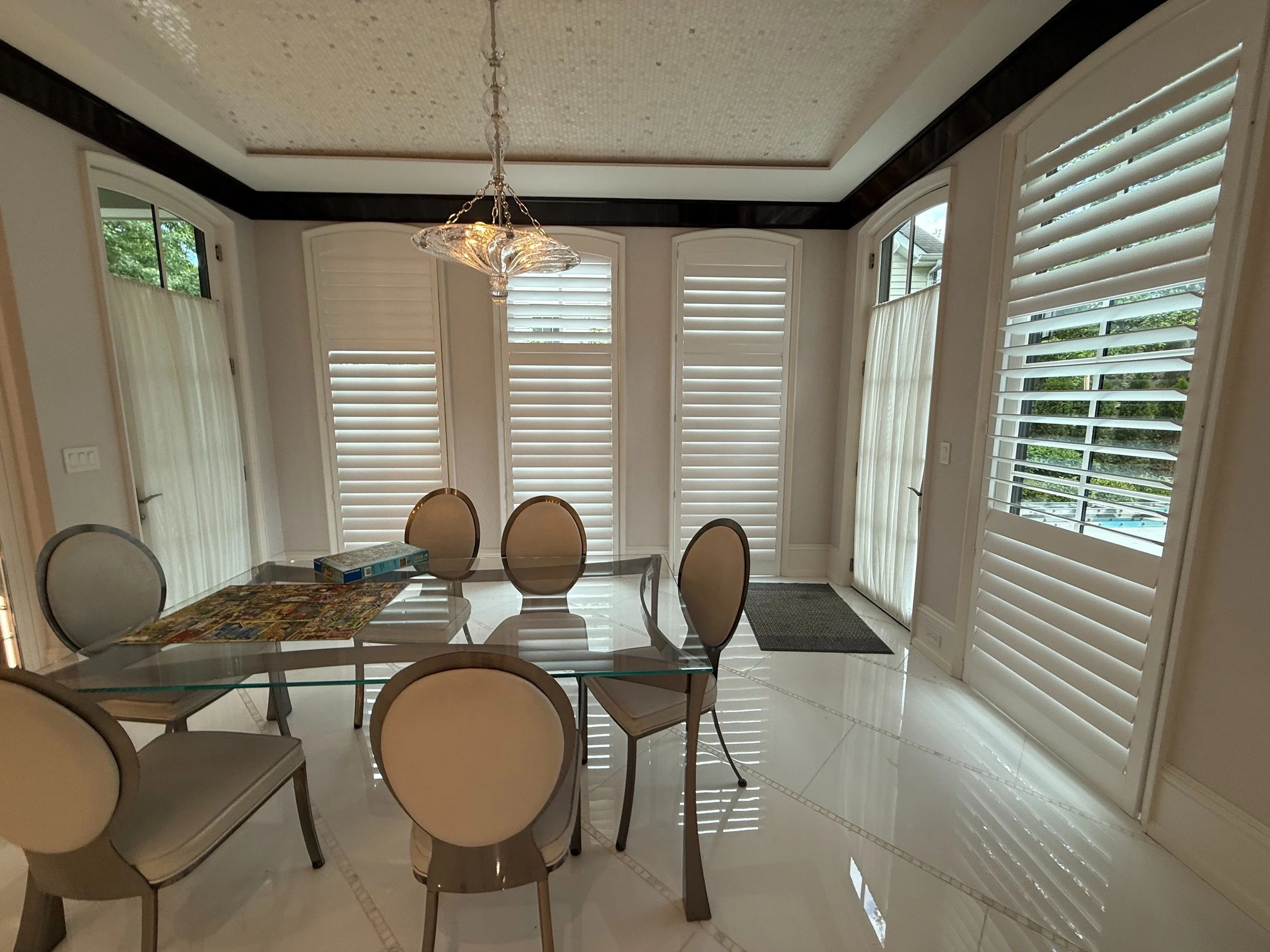 Elegant breakfast room with custom plantation shutters on arched bay windows, glass table, crystal chandelier, NYC