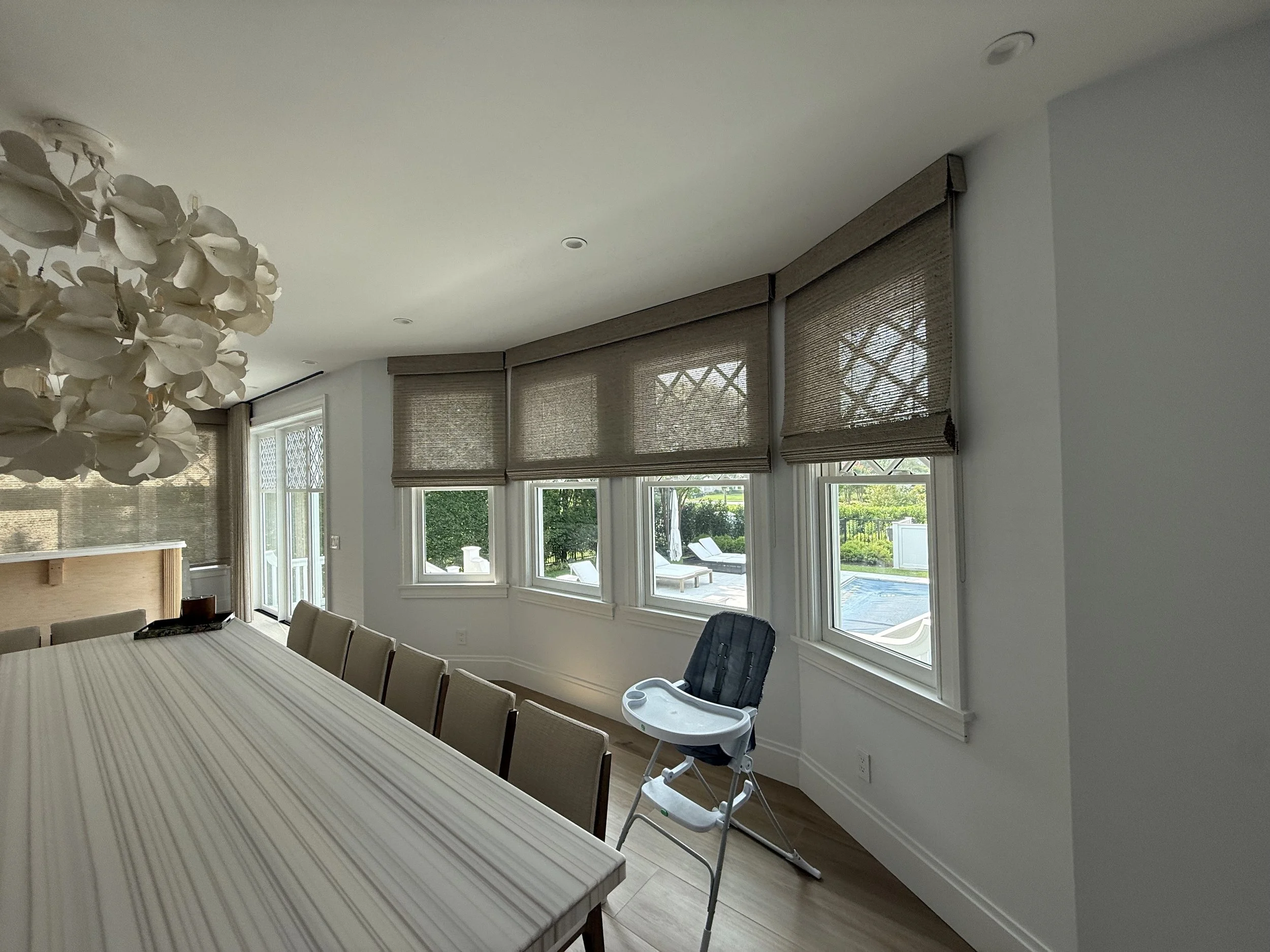 Dining room with multiple woven roman shades on curved window wall and pool views, NYC Rafael Interiors