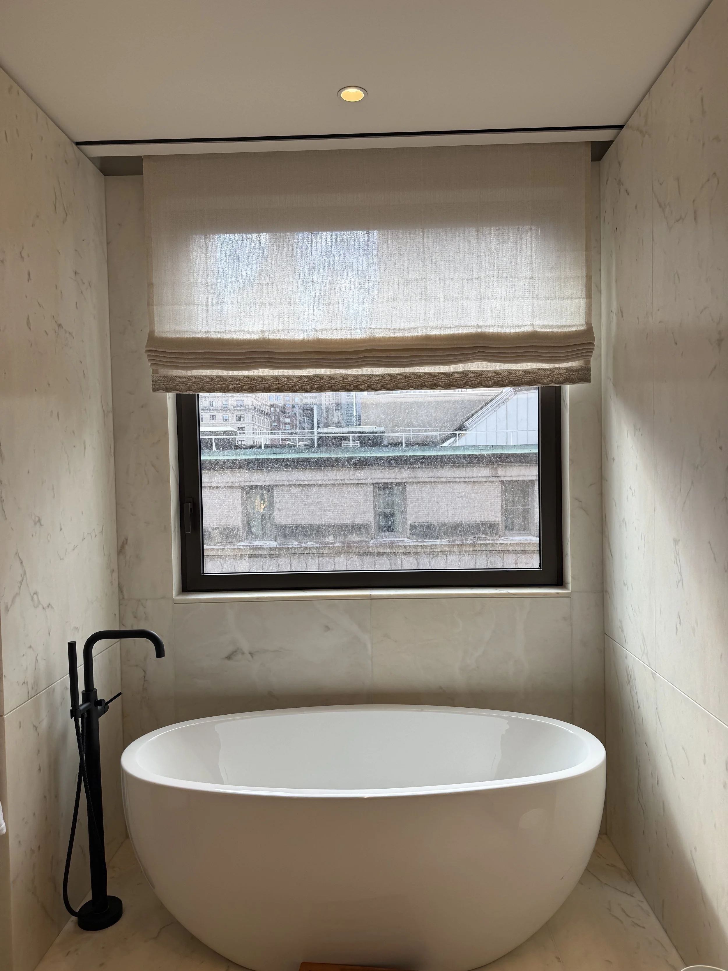 Flat linen roman shade above city view window with freestanding oval soaking tub and marble walls, NYC Rafael Interiors