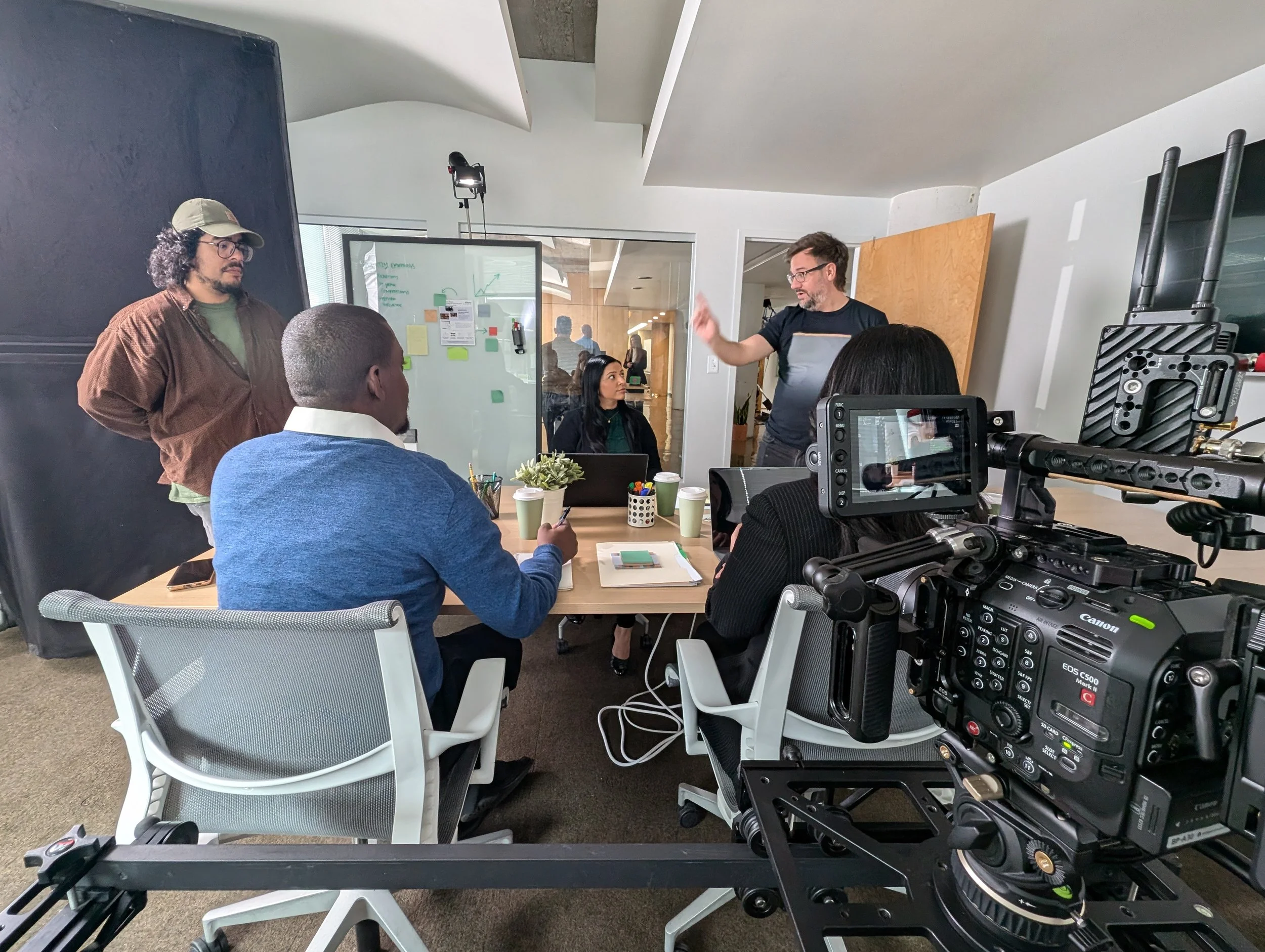 A diverse group of five people engaged in a discussion in a modern office space, with a large video camera recording the scene.