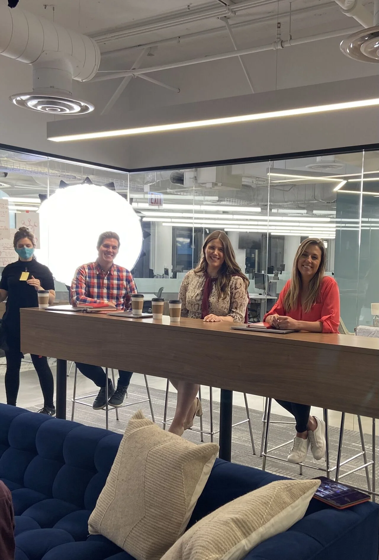 Four people sitting and standing behind a high wooden table in an office space, with three women and one man, three having coffee cups in front of them, in a modern office with glass walls and a white ceiling with visible ductwork.