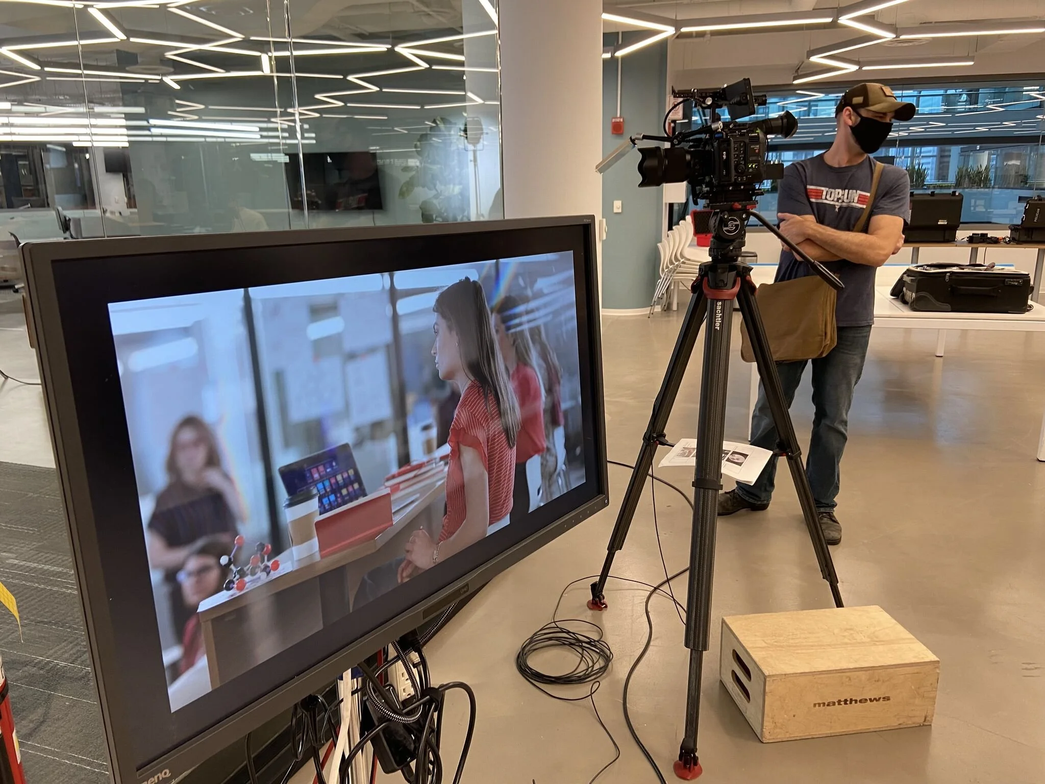 A person operating a camera on a tripod in a studio, filming a woman sitting at a table with a laptop and coffee cup, with the woman's reflection visible on a large monitor in the foreground.