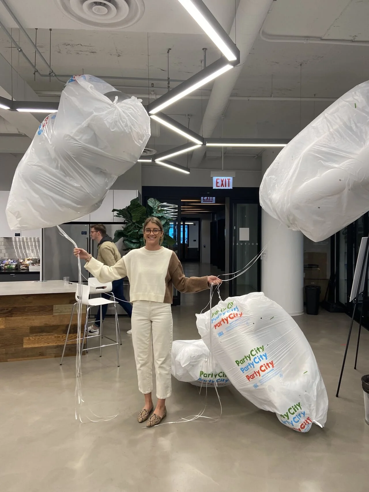 A woman is standing inside a modern indoor space holding two large white balloons and is decorated with multiple big white garbage bags with colorful print that reads "Party City." She is smiling and wearing a cream sweater with brown sleeves and cre