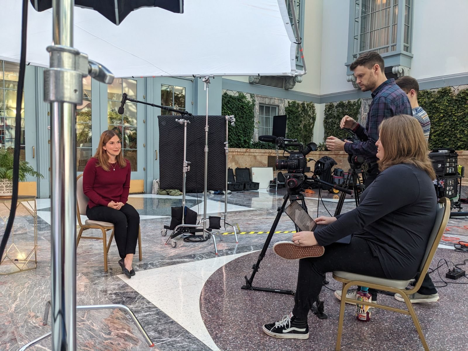 A woman in a maroon sweater and black pants is being interviewed or filmed by a camera crew in a spacious indoor setting with marble flooring and large glass doors. A man in a plaid shirt and a woman in black sit nearby, operating camera equipment an