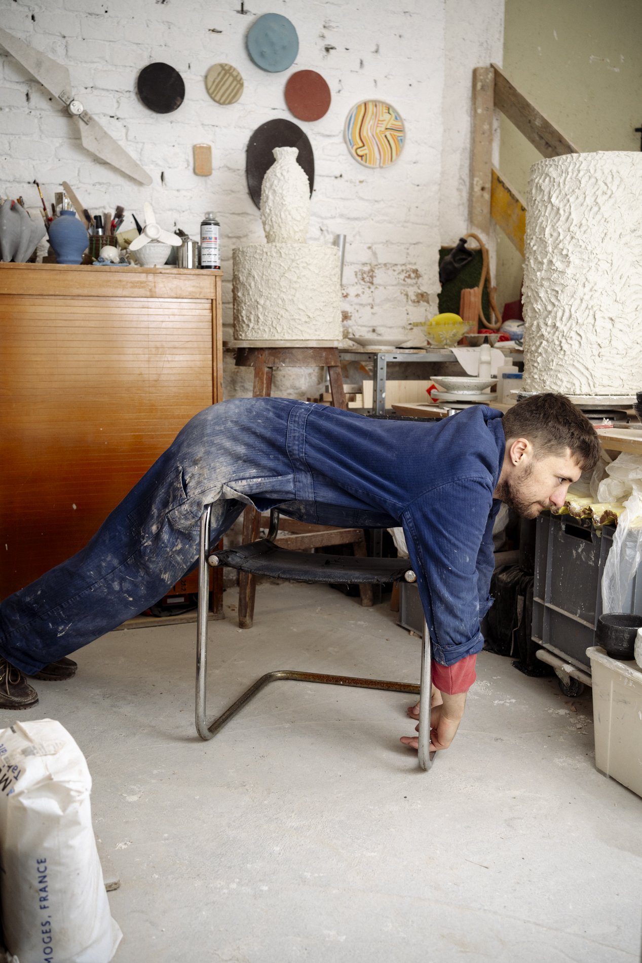 Ein Mann in blauer Arbeitskleidung macht eine Plank aus Ton in einem Atelier. Der Raum ist mit Kunstwerken, Werkzeugen und Farben ausgestattet.