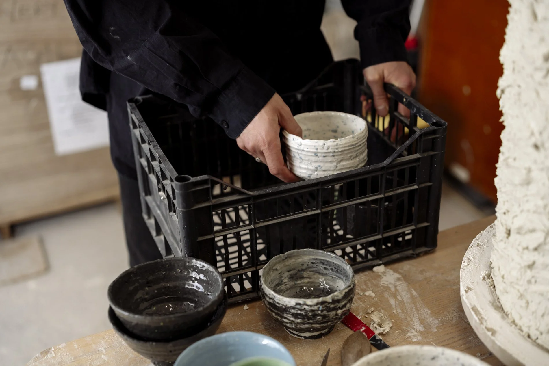 Person beim Keramikhandwerk, hält eine geformte Keramikschale, in einer Werkstatt, umgeben von weiteren Keramikgefäßen auf einem Tisch.