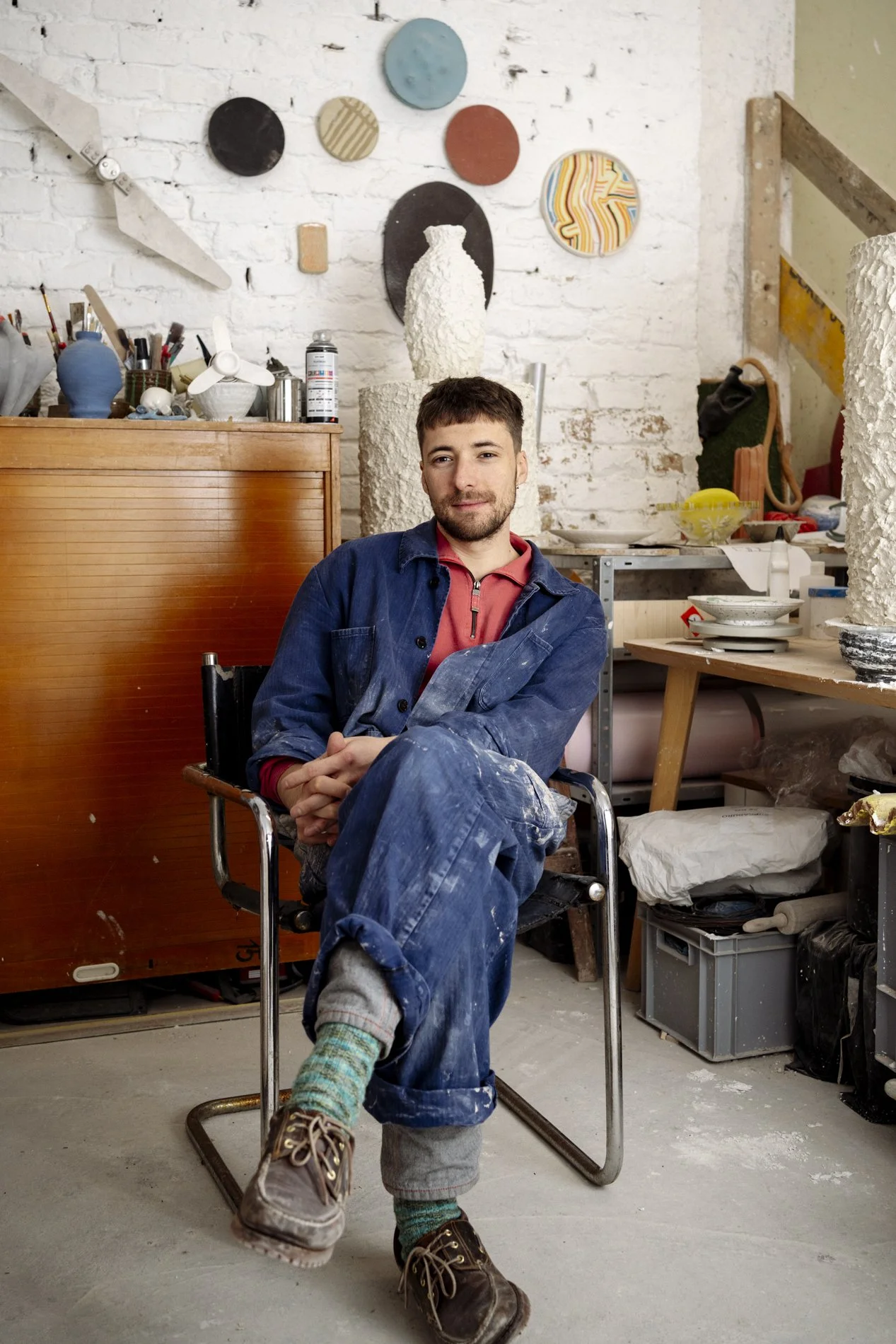Junge Künstler in einer Werkstatt, sitzt auf einem Stuhl mit verschmutzten blauen Berufskleidung, im Hintergrund sind Wandkunst, Werkzeuge, Skulpturen und Arbeitsmaterialien sichtbar.