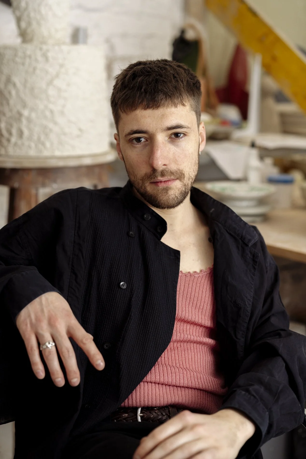 Junger Mann mit dunklem Hemd und rotem Tanktop sitzend, im Hintergrund eine Werkstatt mit Keramik und Werkzeugen.