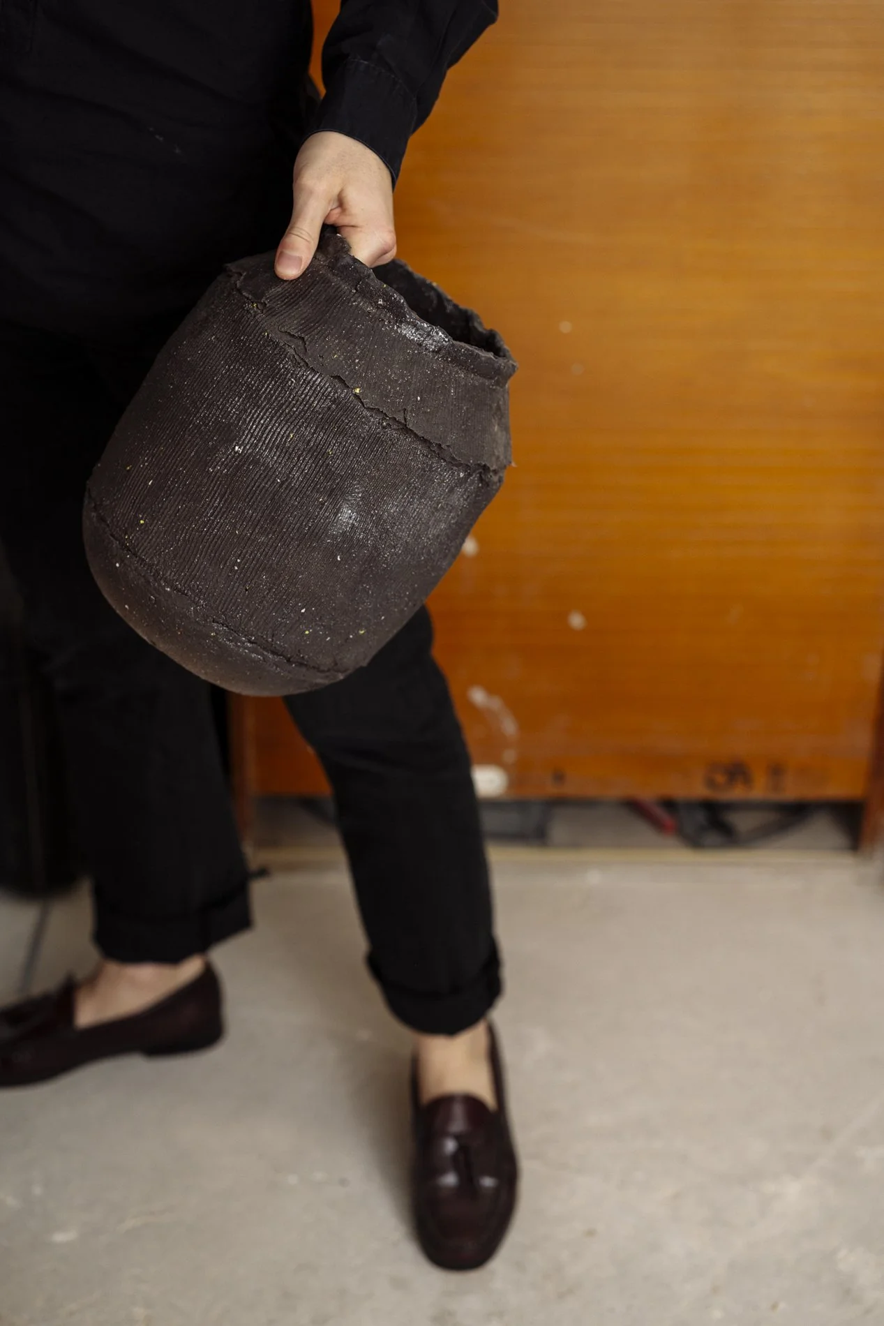 Eine Person hält einen alten, beschädigten, schwarzen Topf, während sie in einem Raum mit Holzwand steht.