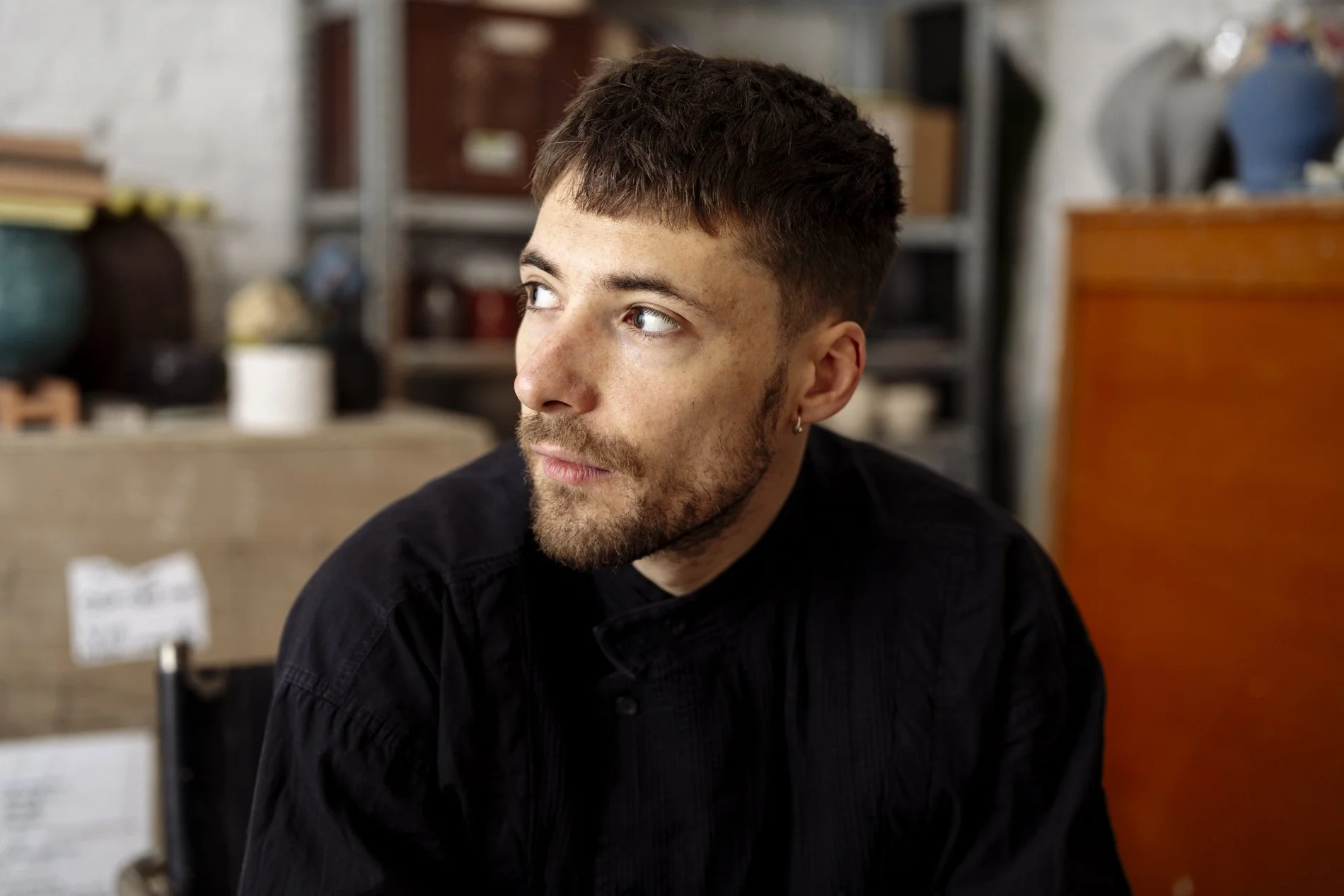 Porträt eines jungen Mannes mit einem schwarzen Hemd, Kurzhaarfrisur und Ohrstecker, lächelnd und nach rechts schauend, im Hintergrund Lagerregale und Kartons in einem Raum.