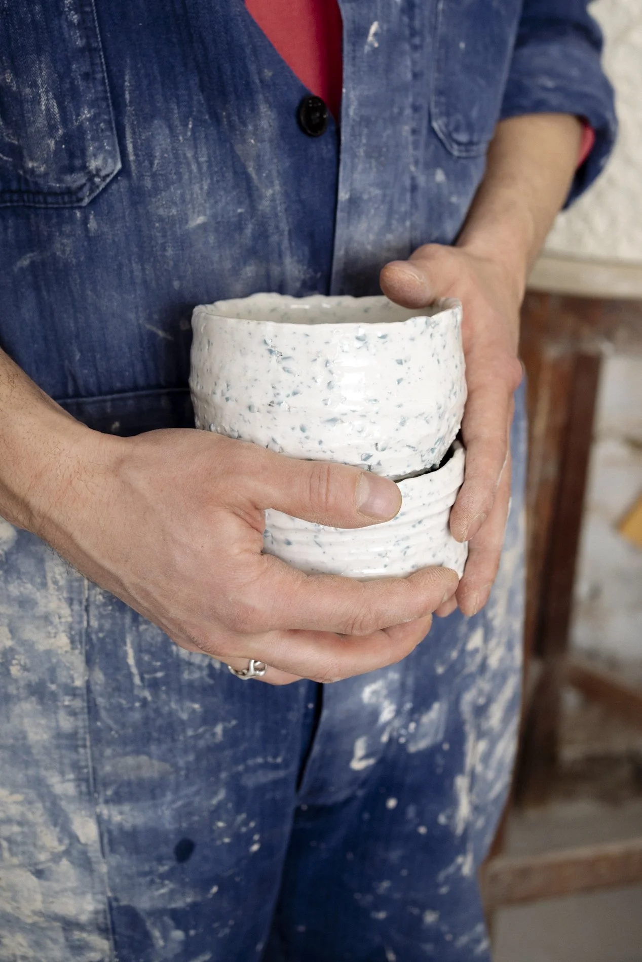 Person hält zwei Tassen keramischer Werkstücke, eine im Vordergrund und eine im Hintergrund, in einer Töpferei, mit blauen Arbeitskleidung, Staub auf der Kleidung.