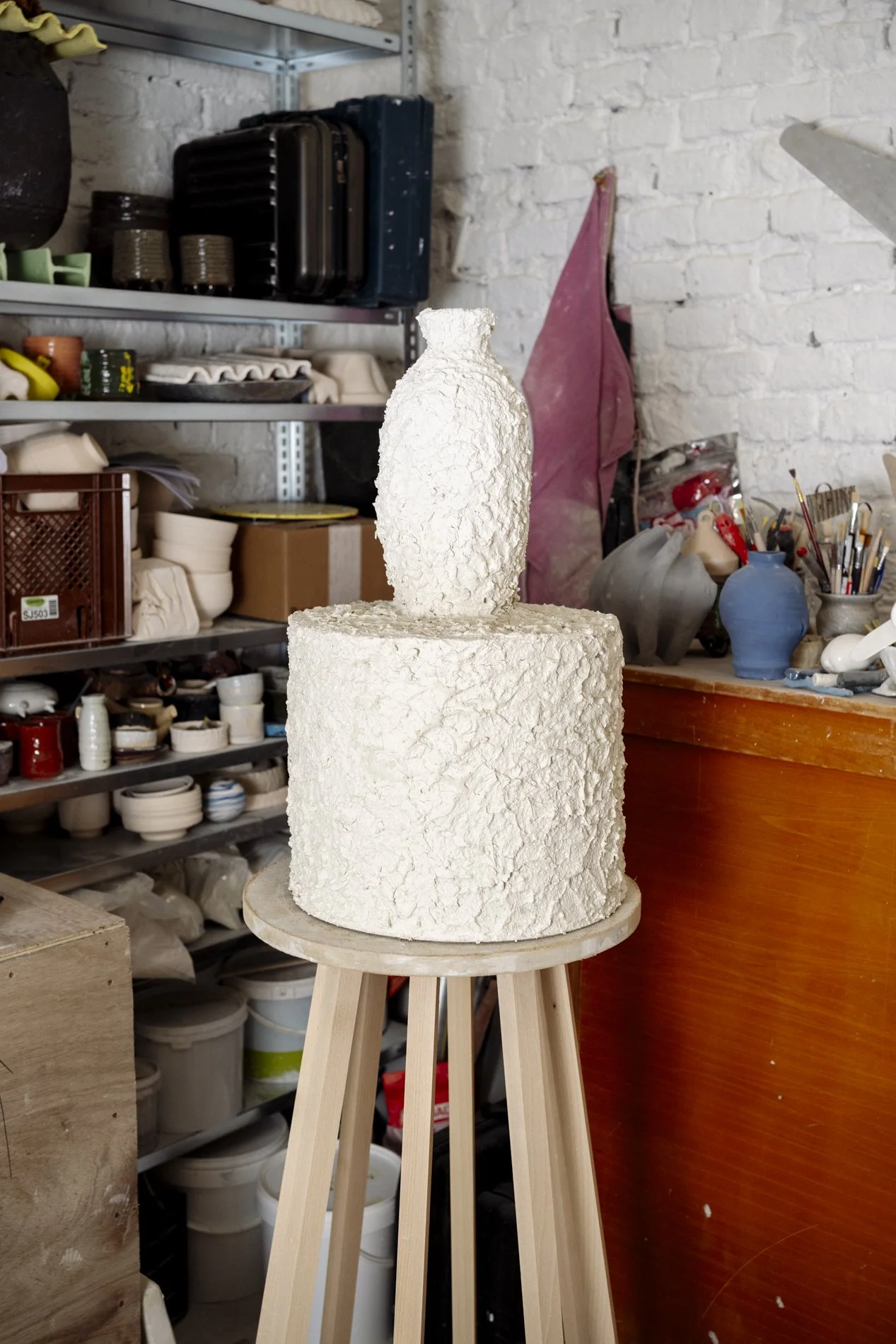 Un unvollendeter Keramikskulptur in Form einer Vase auf einem hohen Holzständer in einem Keramikstudio, umgeben von Regalen mit keramischen Gegenständen und Werkzeugen.