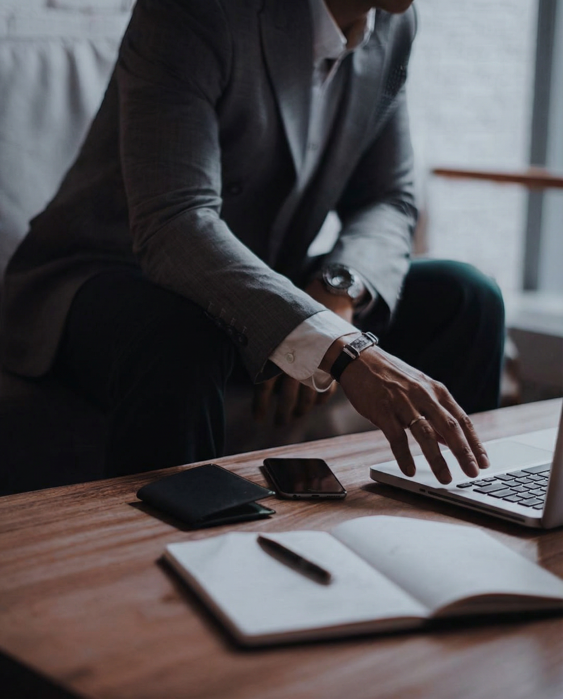A man in a gray blazer working on a laptop at a wooden desk with a notebook, pen, and smartphone.