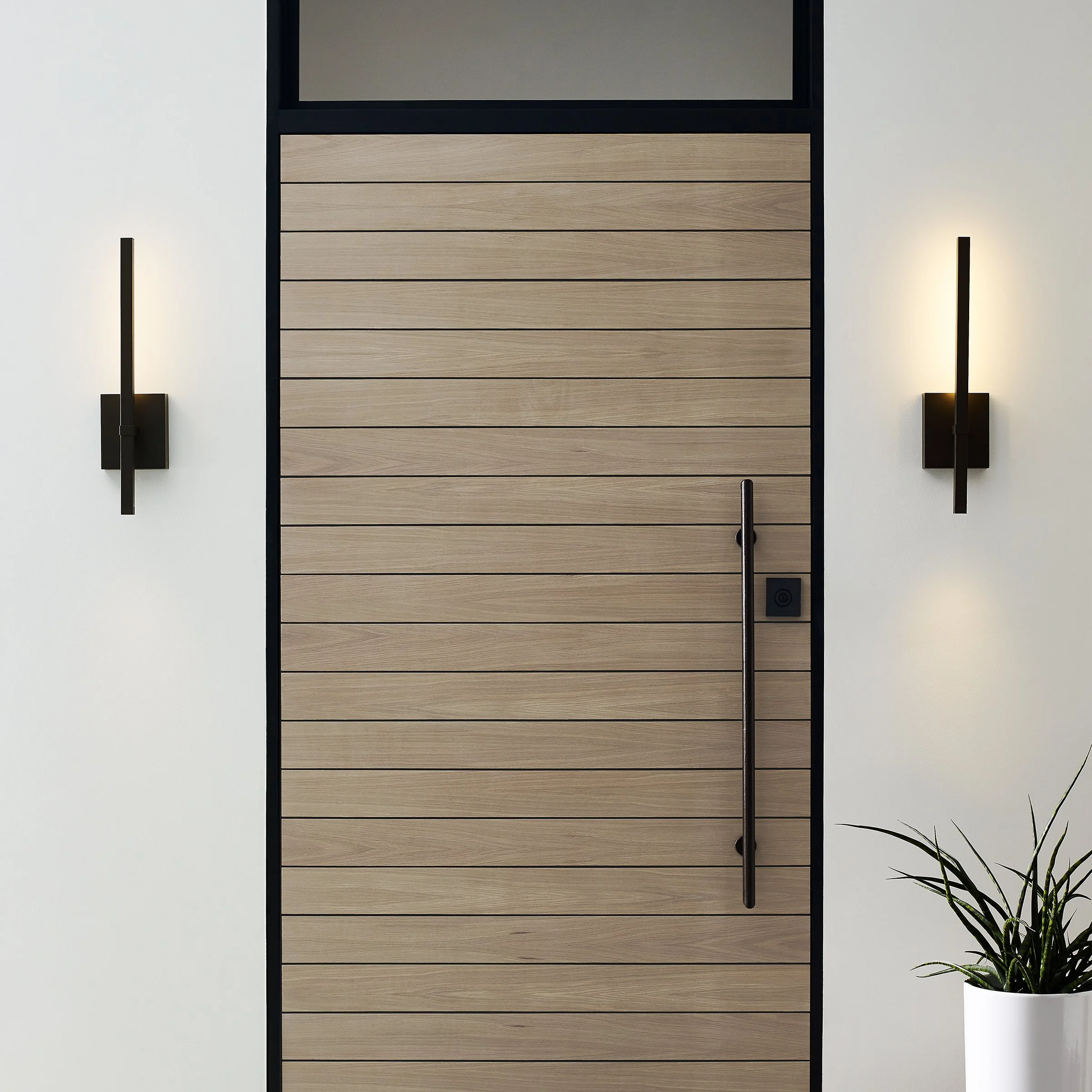 Interior view of a modern room featuring a sliding wooden door with horizontal panels, flanked by two black wall-mounted light fixtures emitting warm light, and a potted leafy green plant in a white pot on the right side.