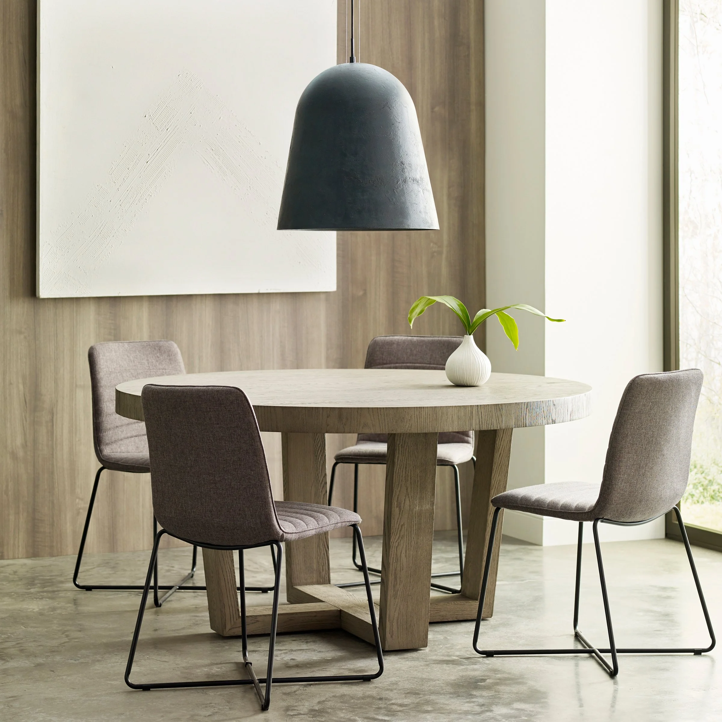 A dining area with a round table, four chairs, a white vase with green leaves, a black pendant light, a textured white art piece, and wooden wall paneling near a window.
