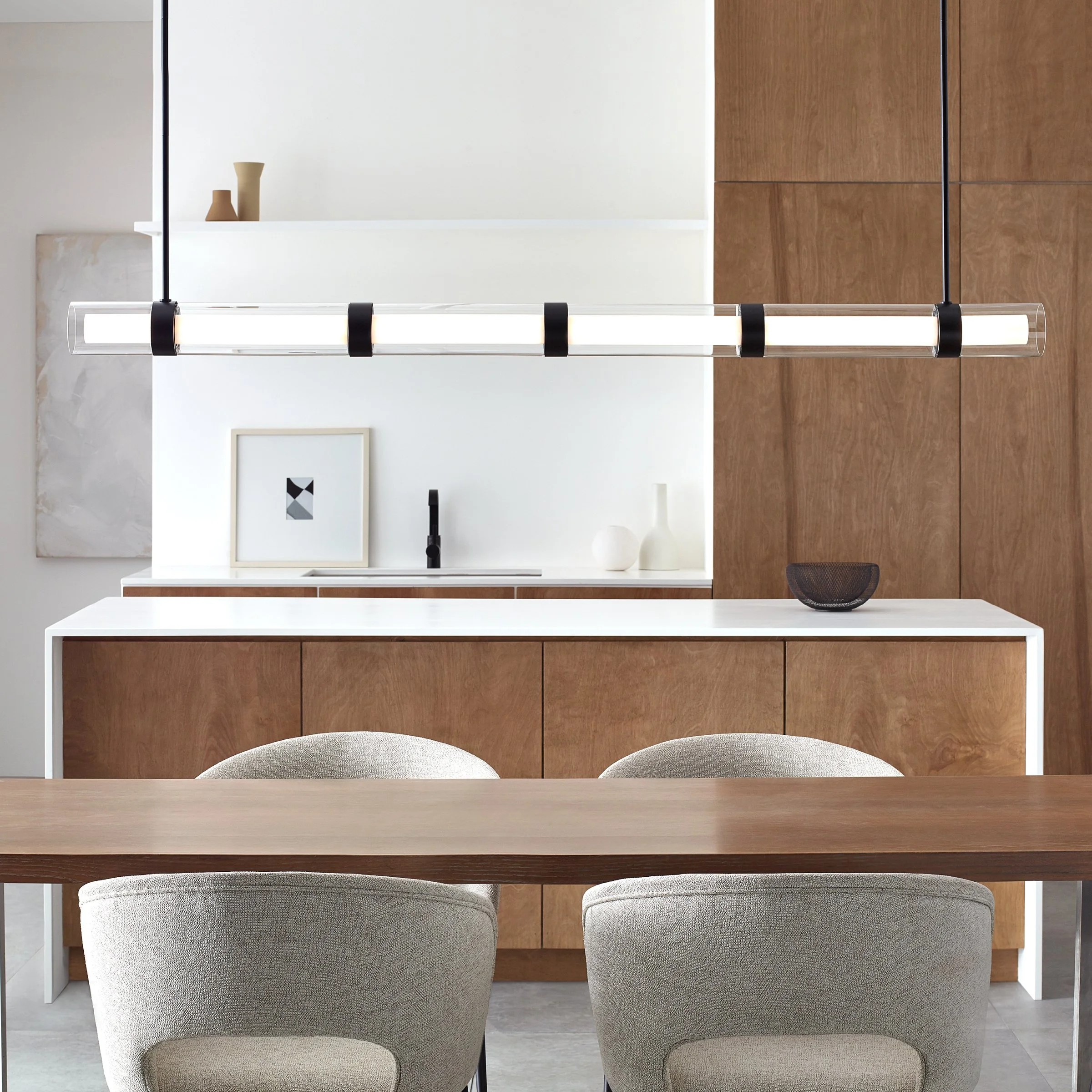 A dining area with a wooden table, two beige upholstered chairs facing the table, a modern hanging LED light fixture, and a wall with a framed artwork. The room has a modern design with minimalist decor.