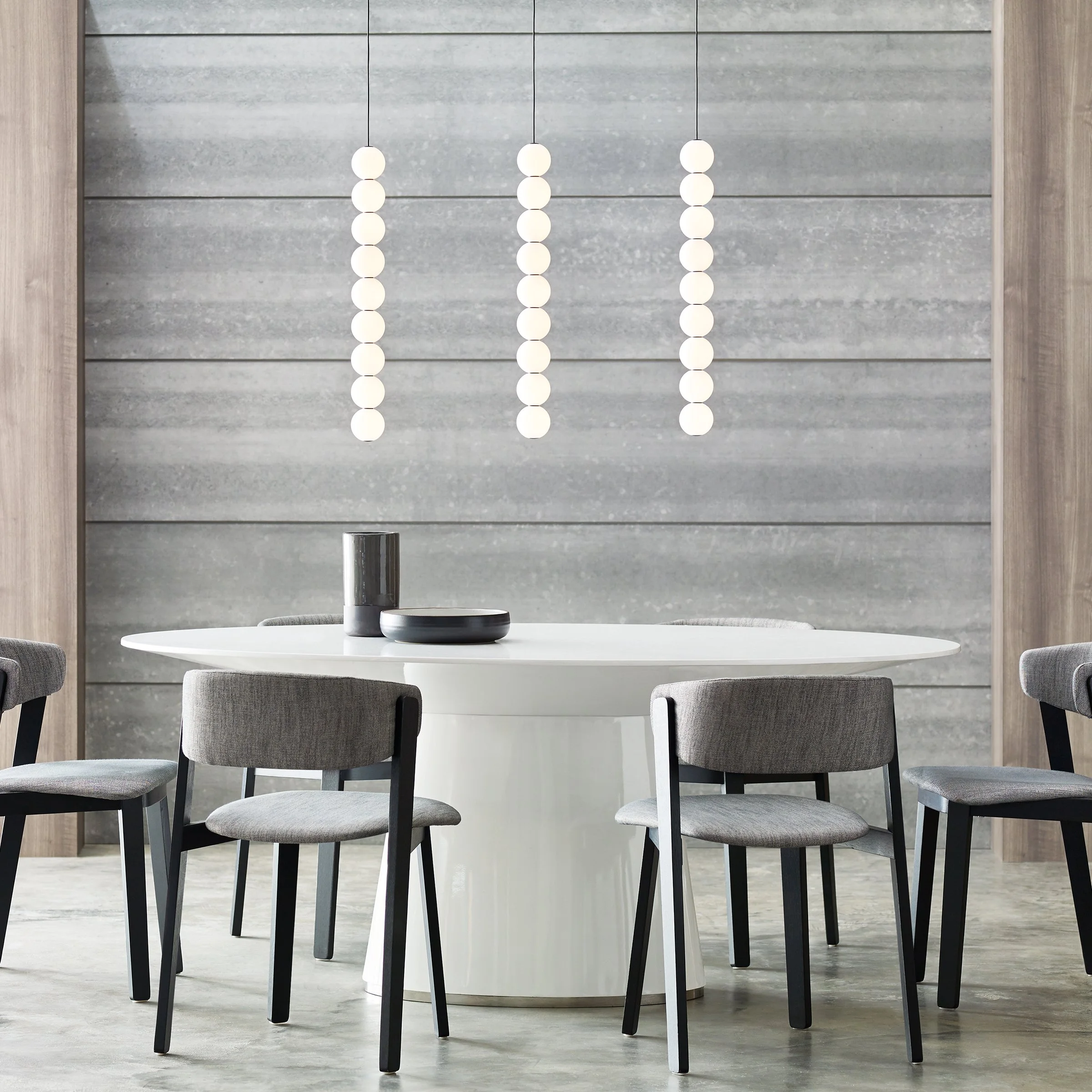 Modern dining or conference room with a round white table, gray upholstered chairs, decorative suspended pendant light fixtures against a gray textured wall.