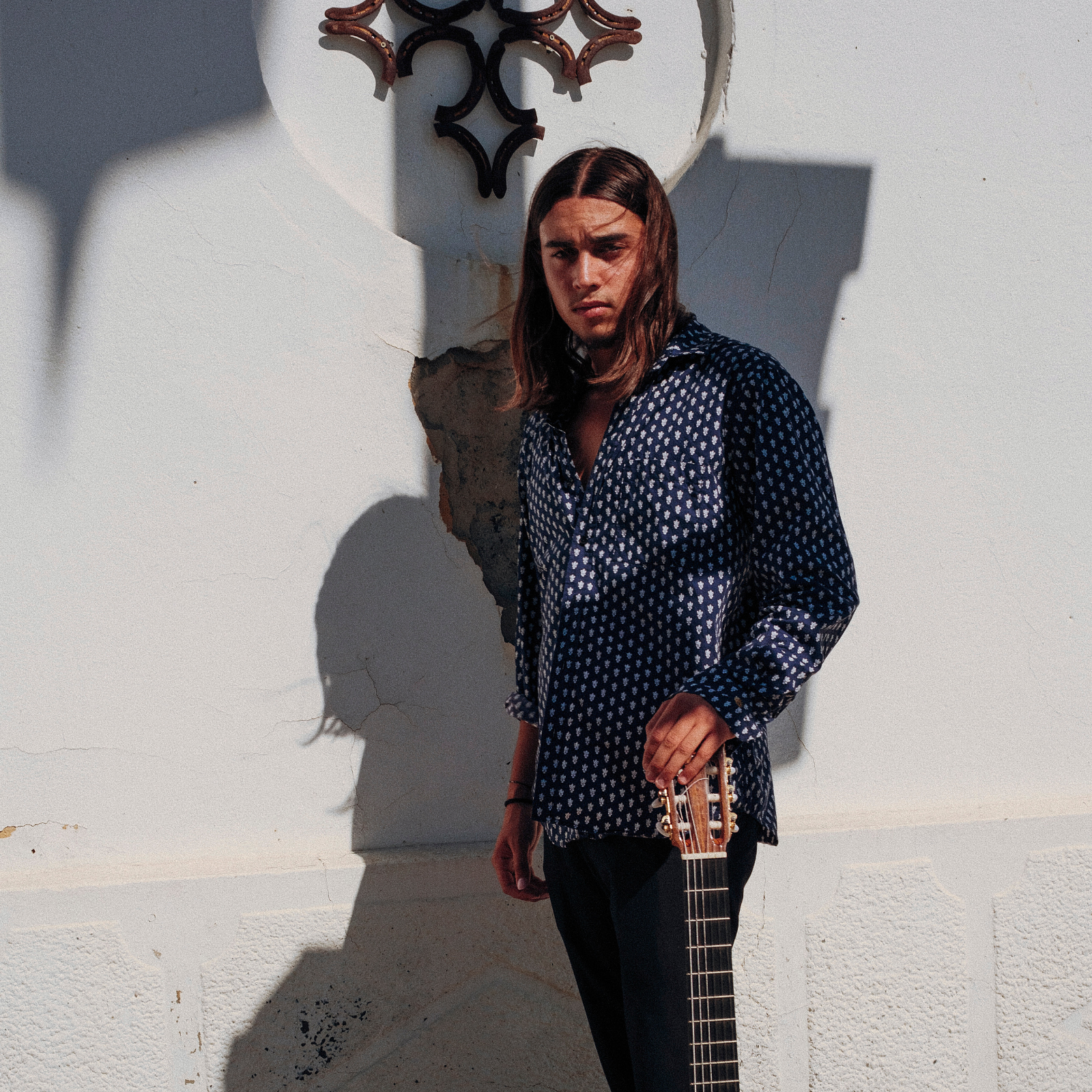 Un jeune homme avec des cheveux longs, portant une chemise à motifs, tient une guitare et se tient devant un mur blanc avec une décoration métallique. Il y a une ombre portée sur le mur.