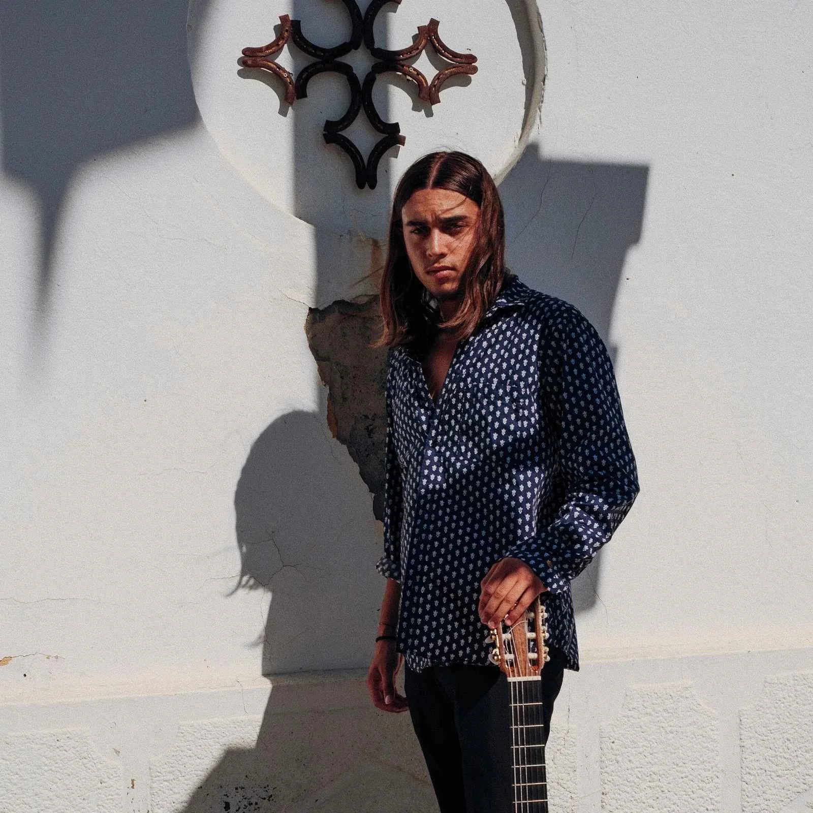 Jeune homme avec des cheveux longs, portant une chemise à pois, tenant une guitare, se tenant devant un mur blanc avec une sculpture en fer noir et cuivre, et son ombre est projetée sur le mur.