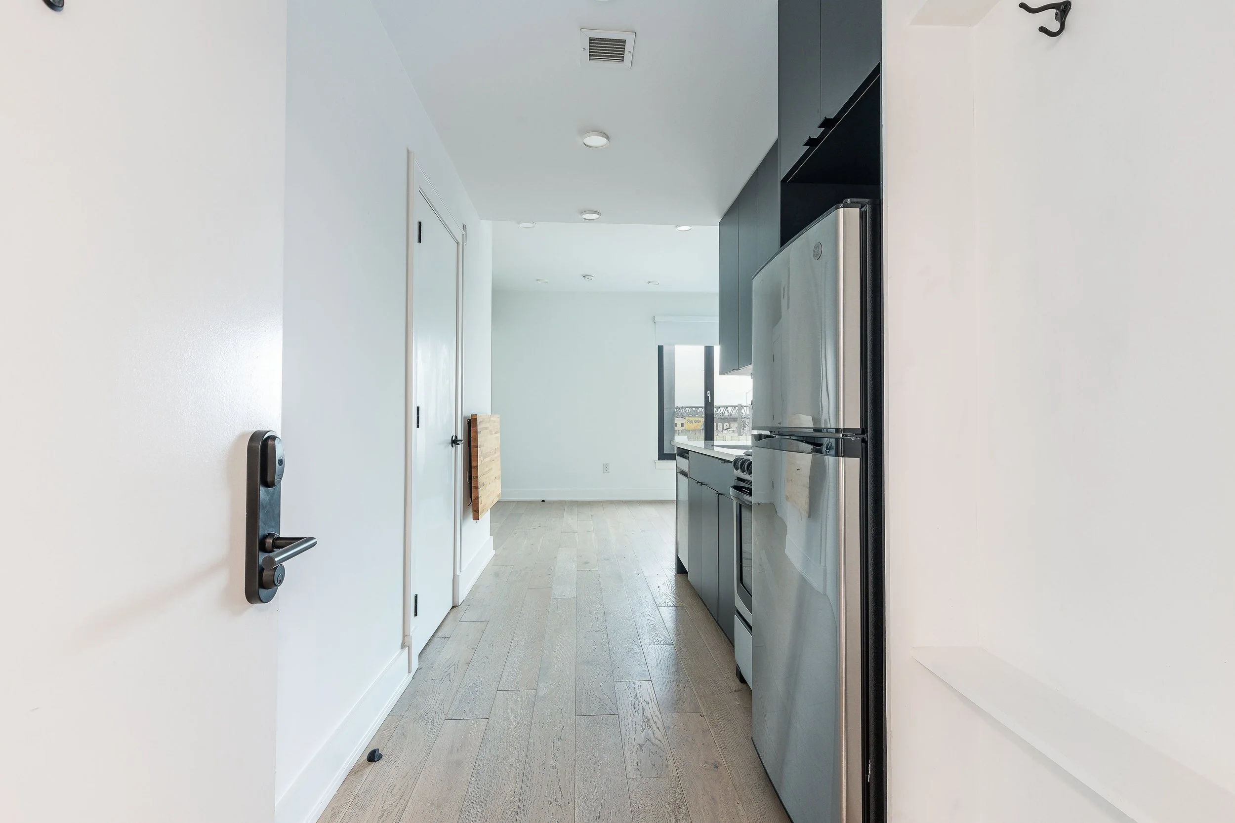 Interior view of a modern kitchen with white walls, wooden flooring, stainless steel refrigerator, black cabinetry, and a window at the end of the room.