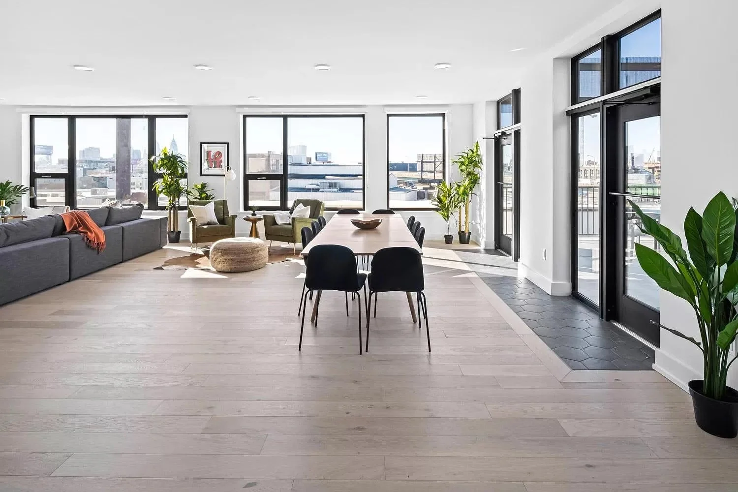 Bright, modern living room and dining area with large windows, green plants, a gray sofa, green chairs, a wooden dining table with black chairs, and city views.