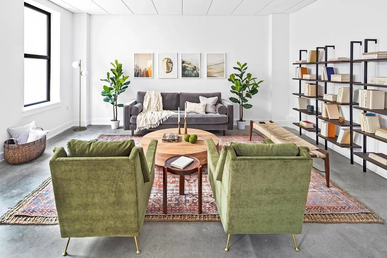 Modern living room with two green armchairs, a gray sofa, a wooden coffee table with candles and decorative items, a rug, a tall black bookshelf filled with books, and potted plants near large windows.