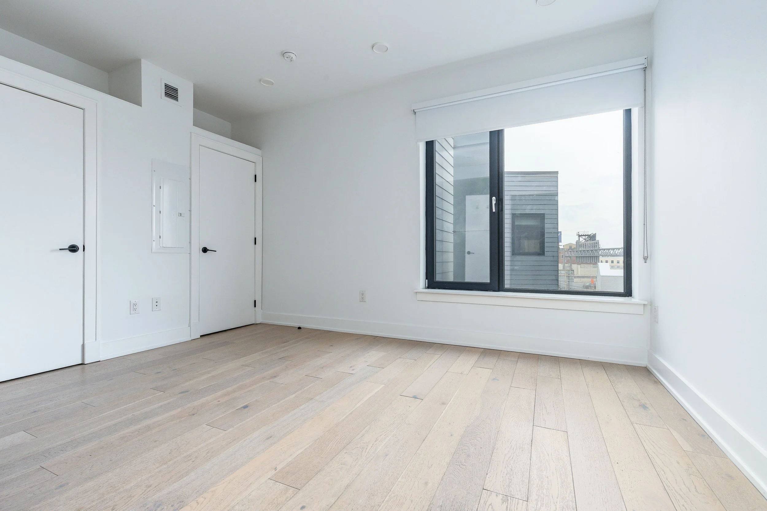 Empty room with light-colored hardwood floors, white walls, a large window with a white blind, and two closed white doors.
