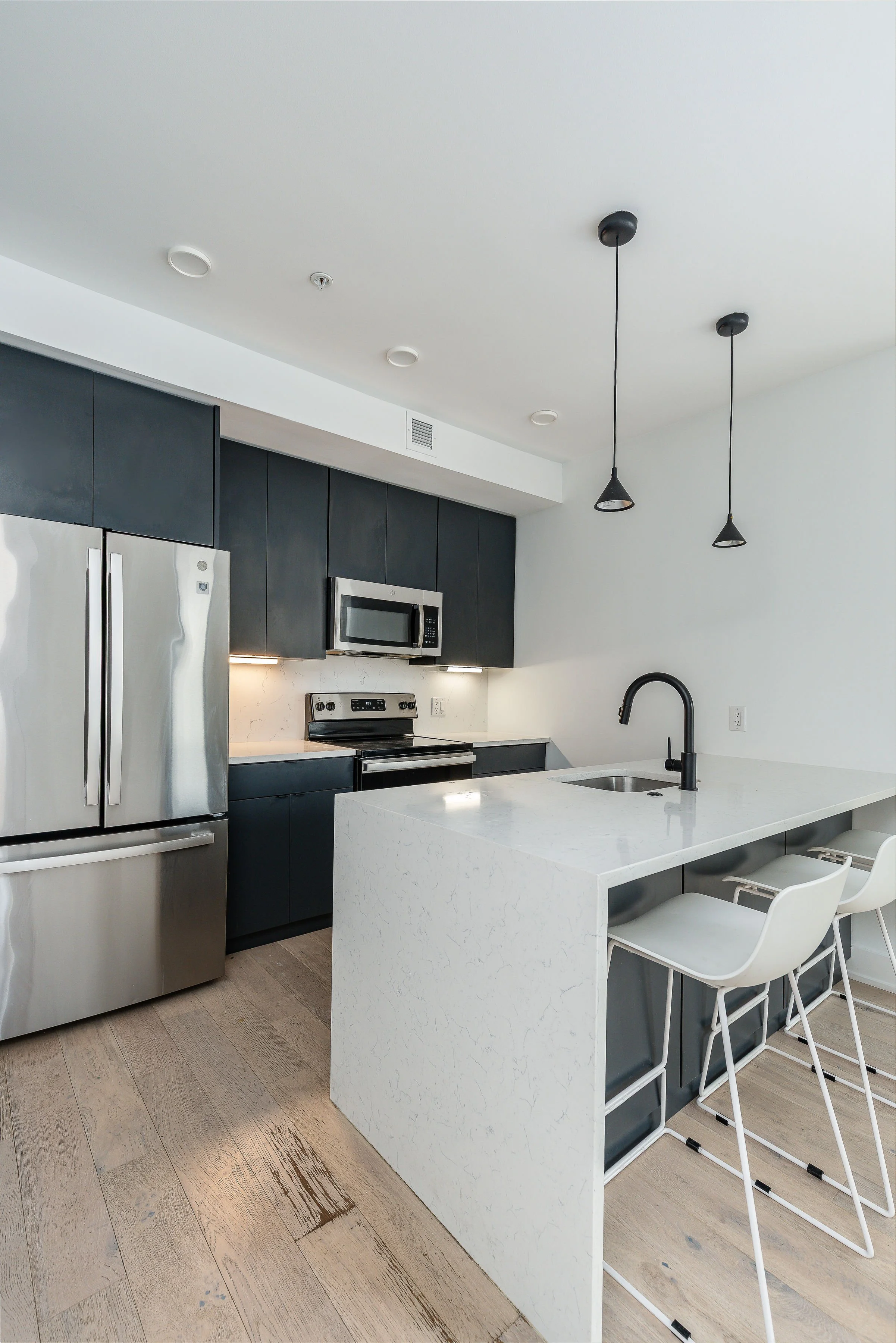 Modern kitchen with white island, black sink faucet, dark upper cabinets, stainless steel refrigerator, microwave, oven, and two white bar stools.